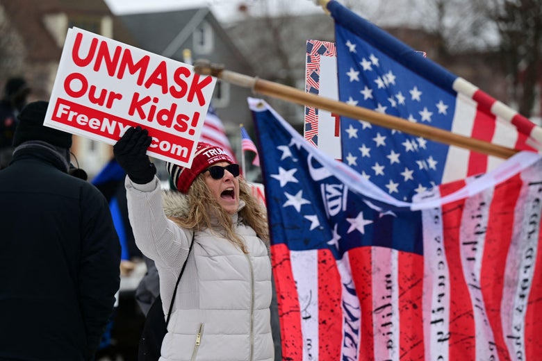 Protester shouting and holding a sign that says "Unmask our kids!" next to American flags with writing on them.