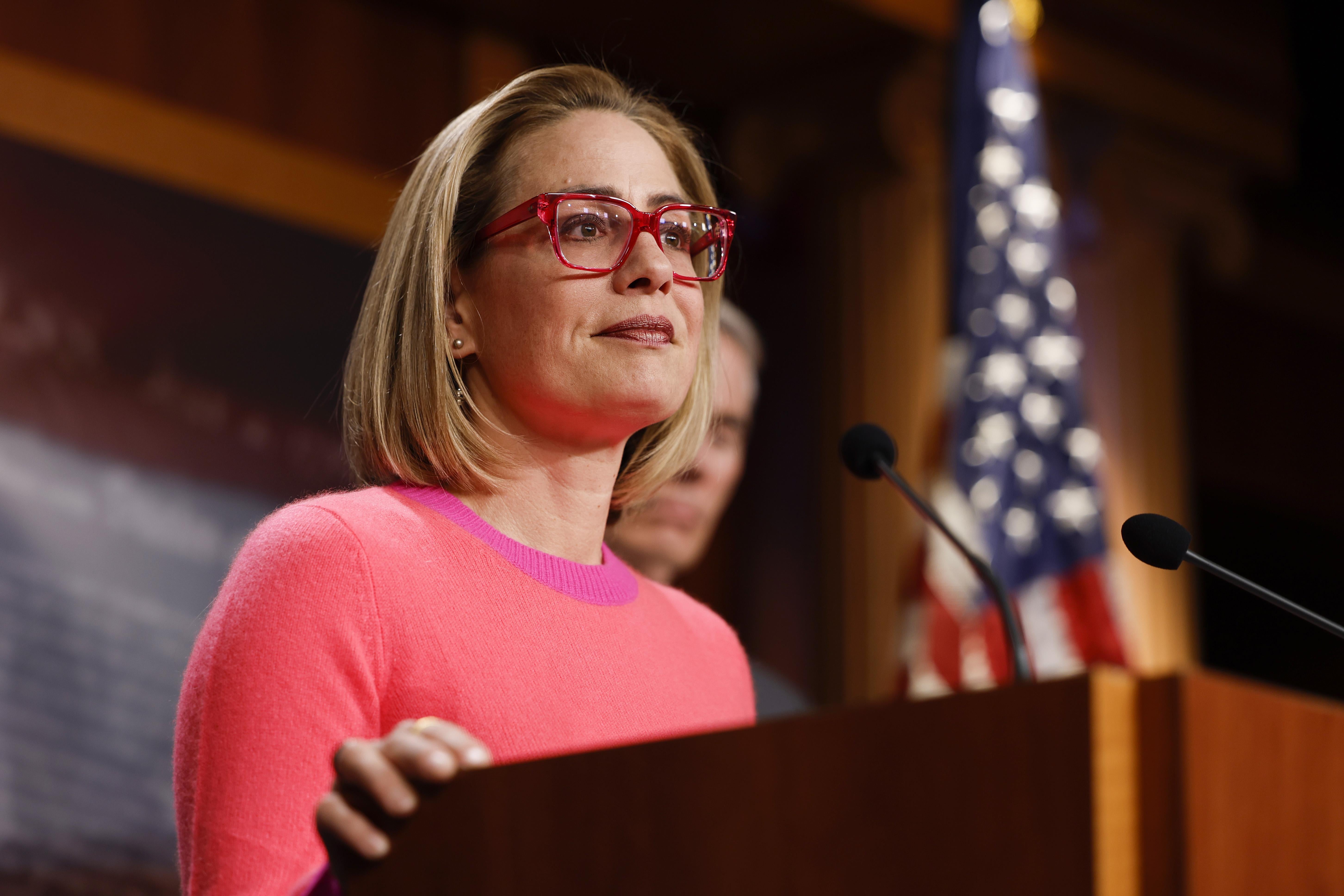 Sinema, wearing a pink sweater and red glasses, stands at a podium.