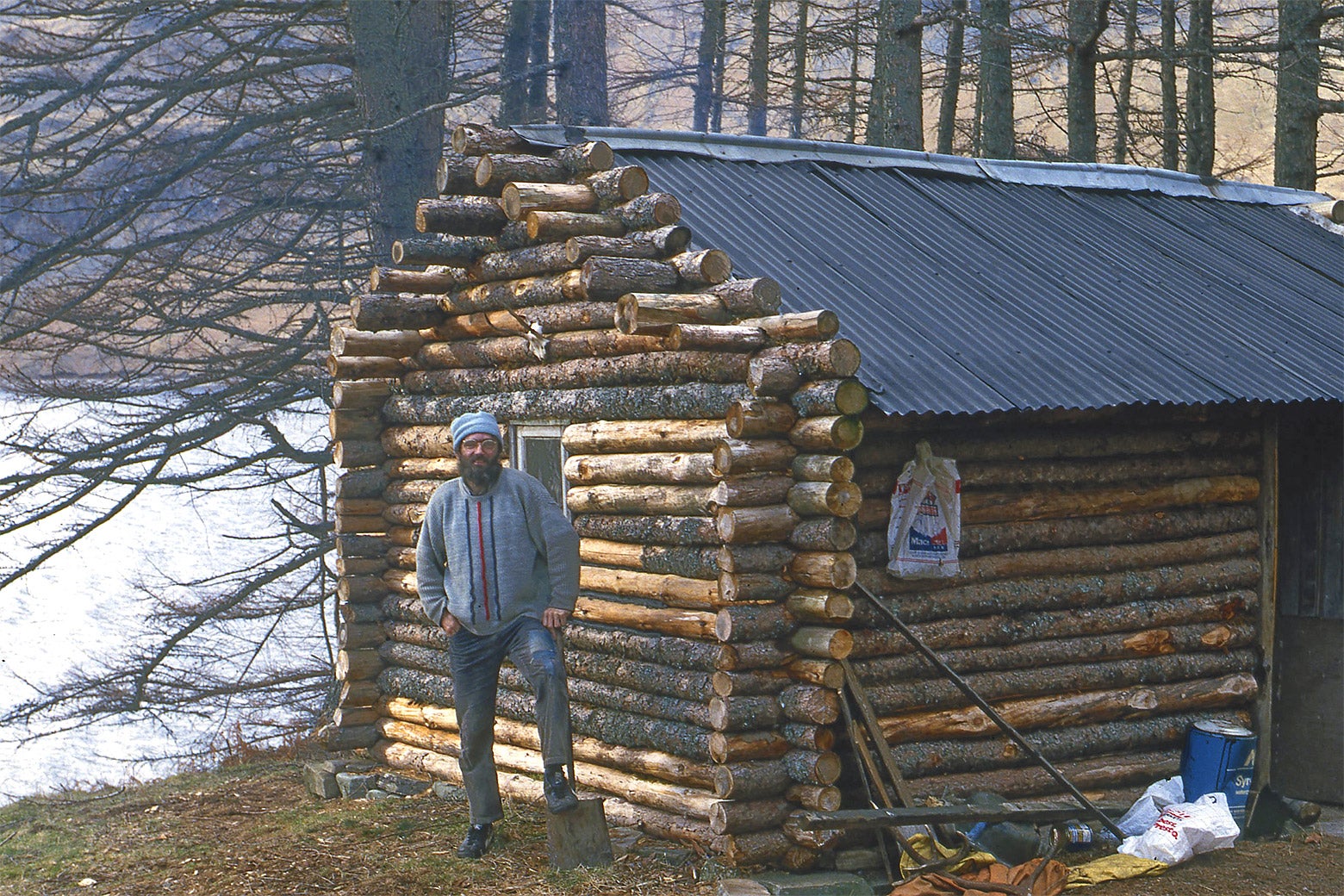 A bearded white man with dark hair stands in front of a log cabin in the woods.