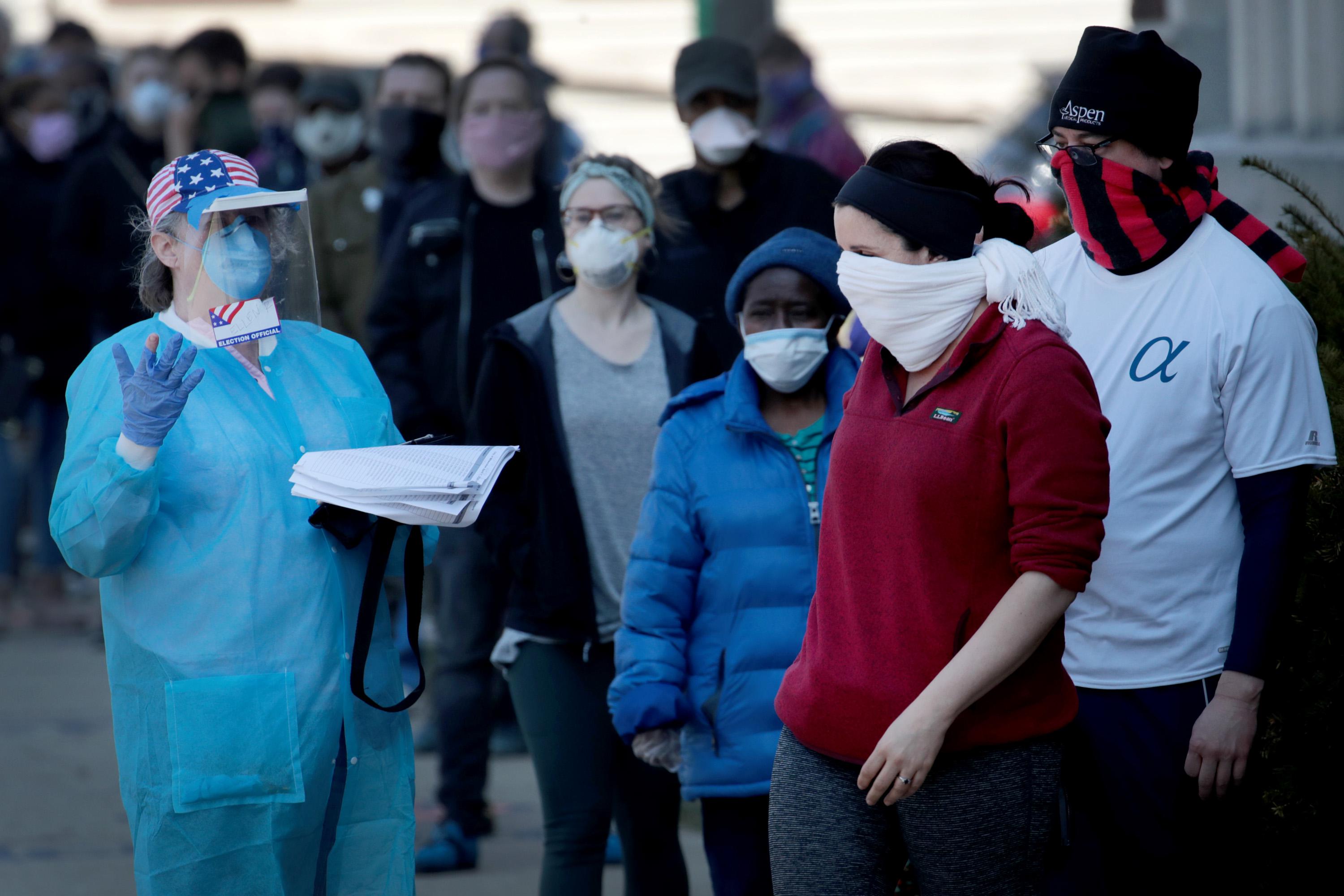 A polling official wearing a gown, gloves, and a face mask directs people in line wearing cloth face coverings