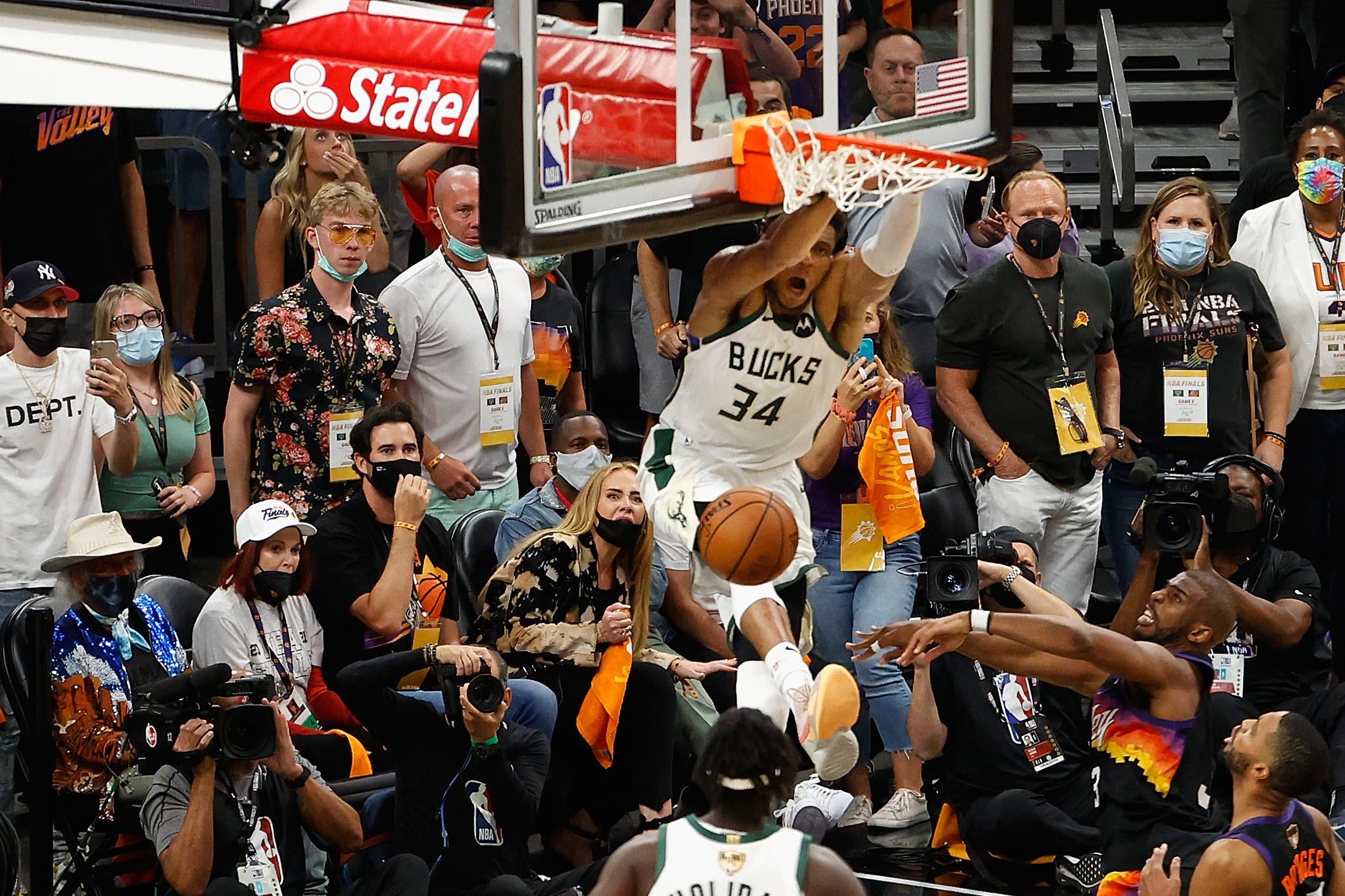 Giannis dunks on the side of the hoop with mouth agape and legs flying as Jrue Holiday, Chris Paul, Mikal Bridges, and Suns fans look on.
