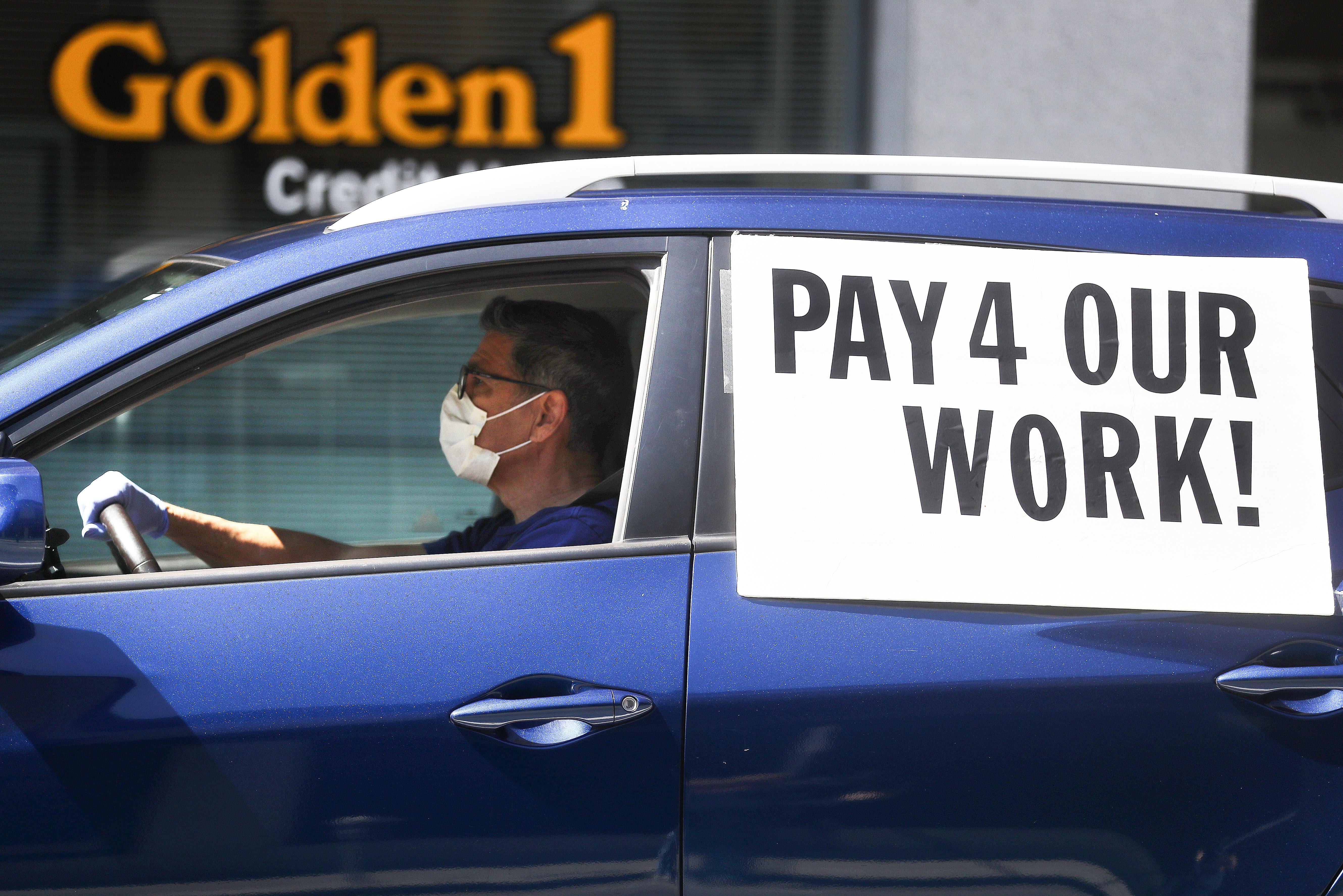 A driver wearing a face mask and gloves, in a car with a sign that says "Pay 4 Our Work!"