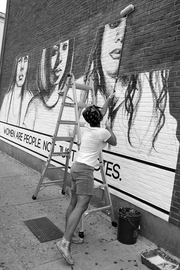 A woman pastes posters on a wall. They feature drawings of women along with their messages to street harassers.  