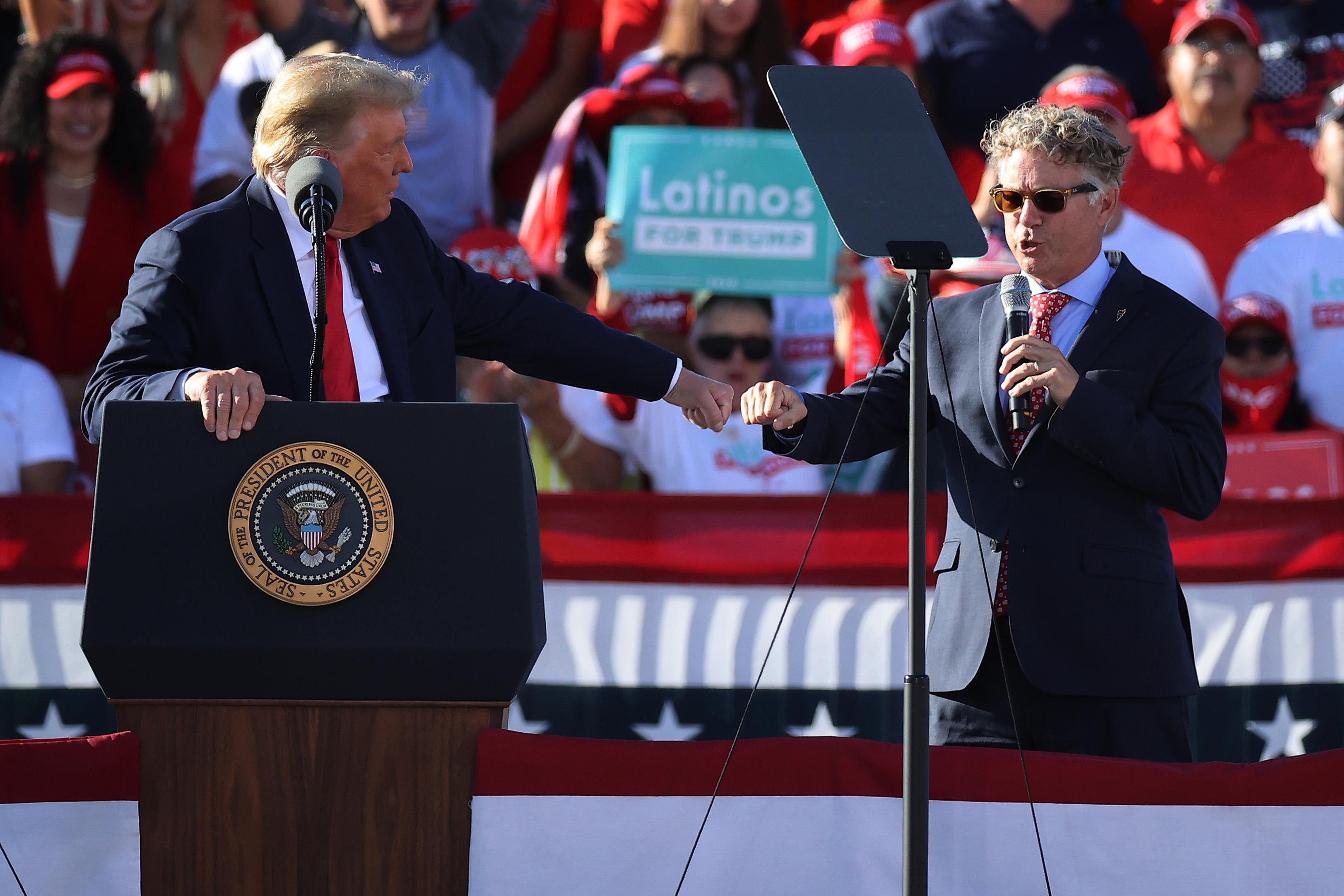 Trump, gripping his podium, fist-bumps Rand Paul as they both stand onstage surrounded by American flag bunting, with a crowd behind them