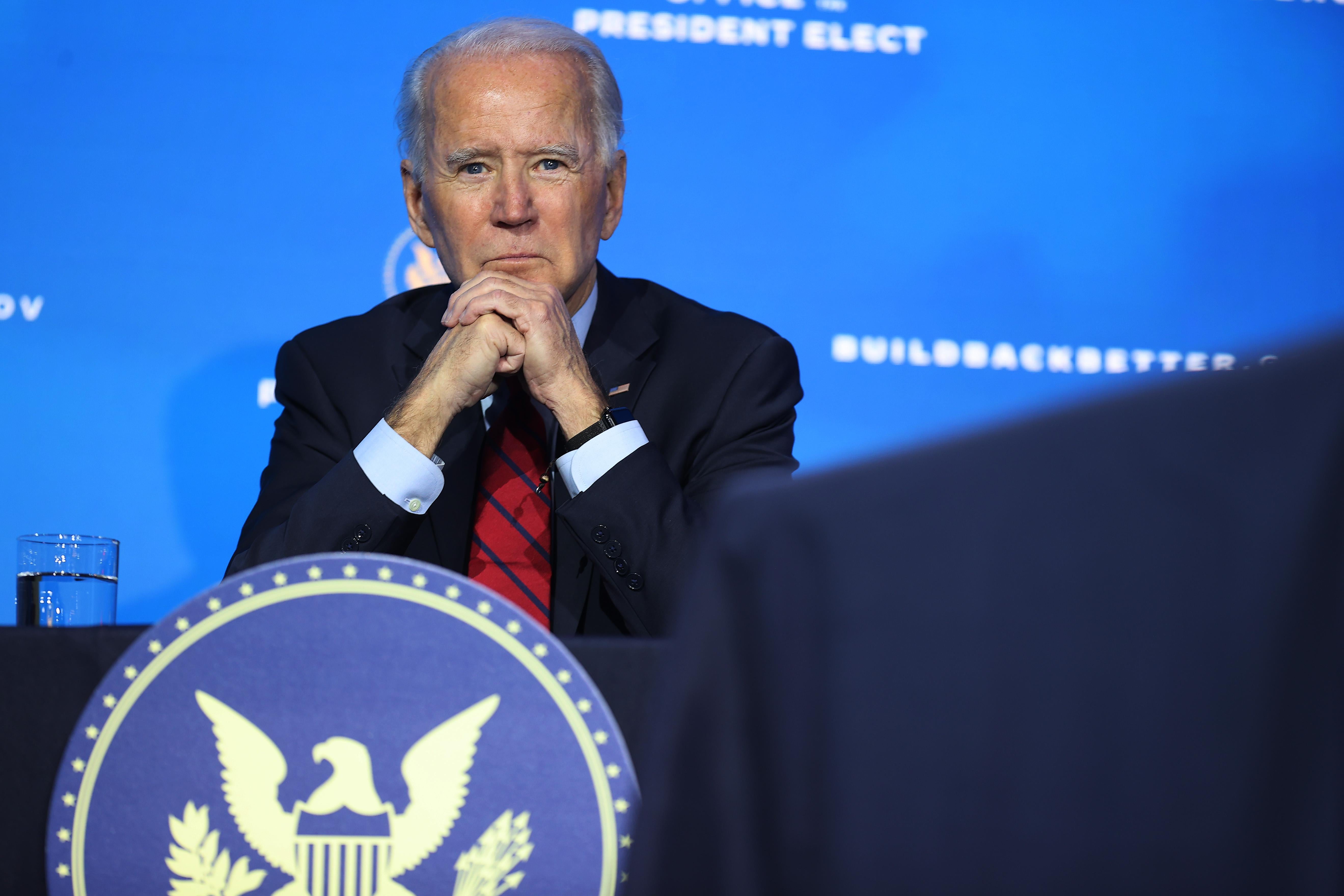 Biden sits with his elbows on a table and his hands clasped, with a presidential seal in front of him