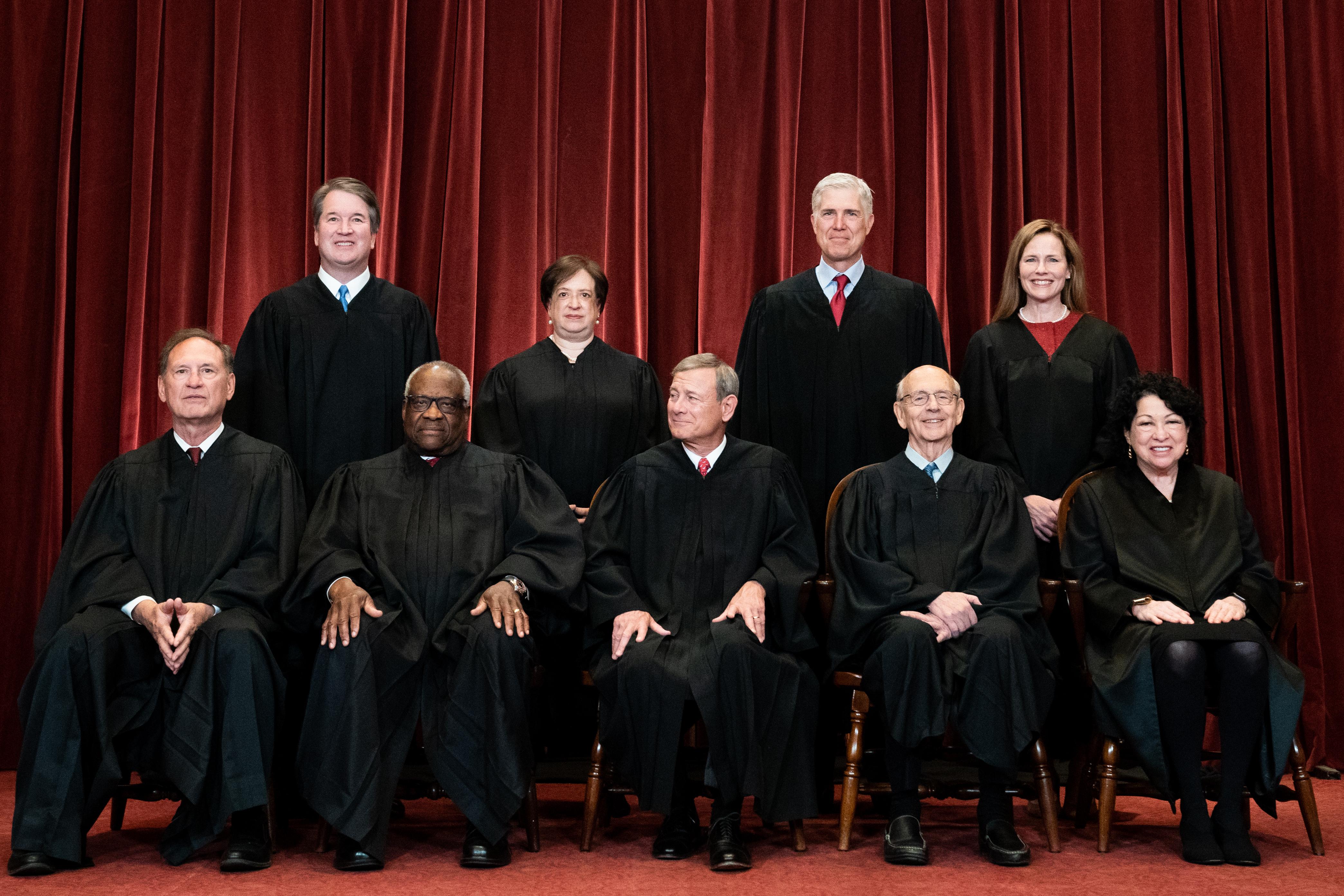 All nine justices of the Supreme Court wearing their robes and smiling in front of a red velvet curtain