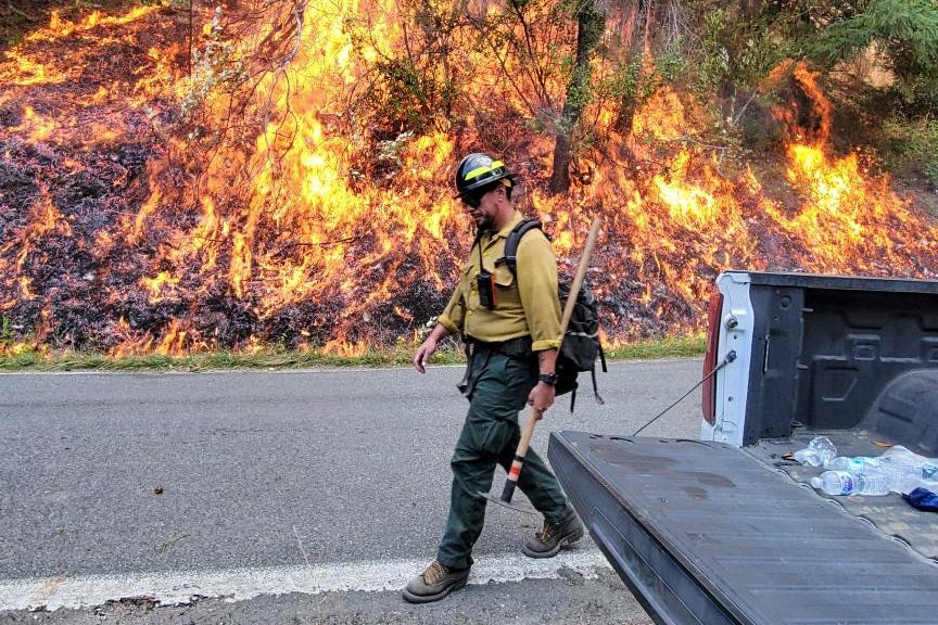 A firefighter in gear walks on a road, past an open pickup truck bed. Behind him, a forest fire burns. 