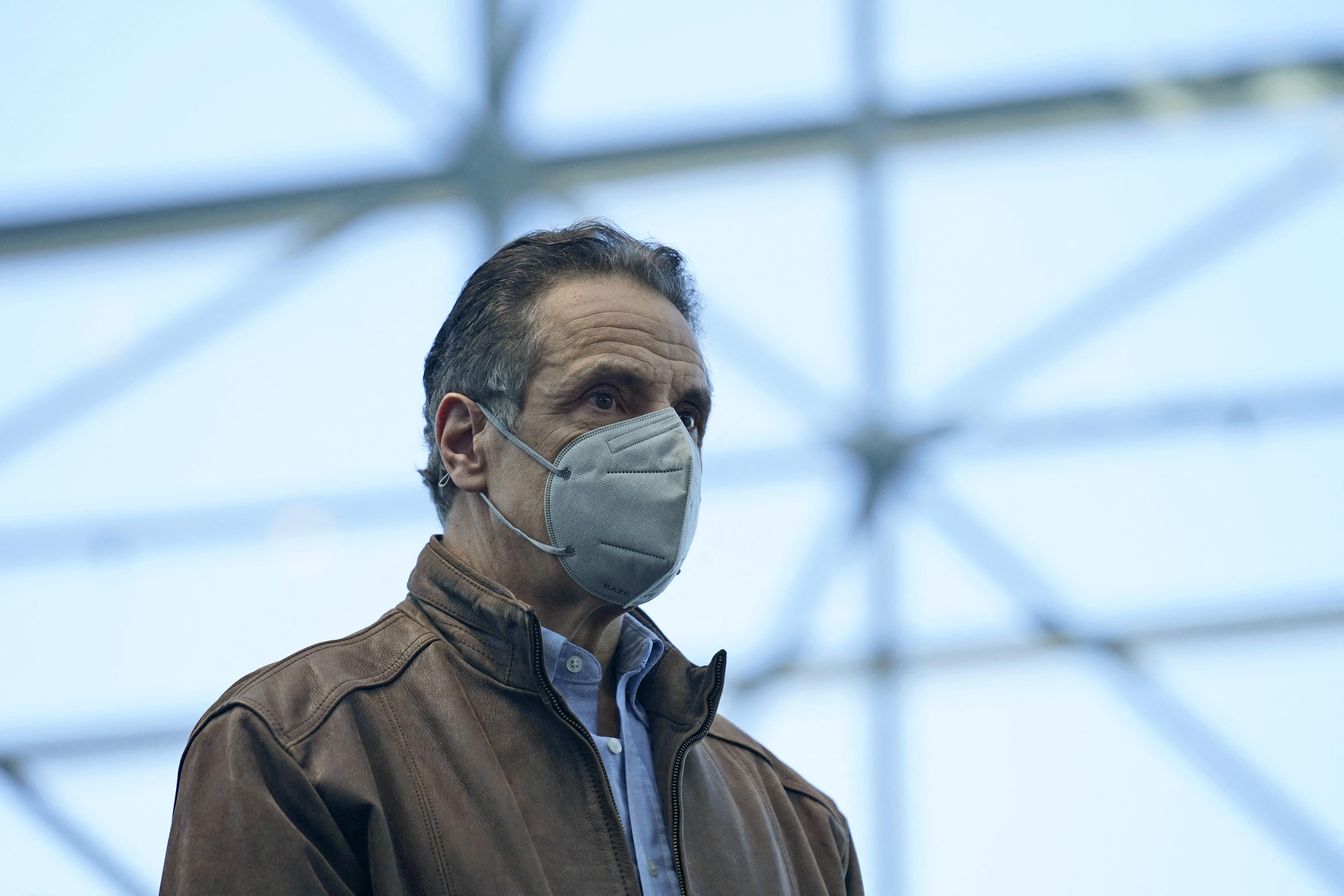 Cuomo stands wearing a gray mask and a brown leather jacket
