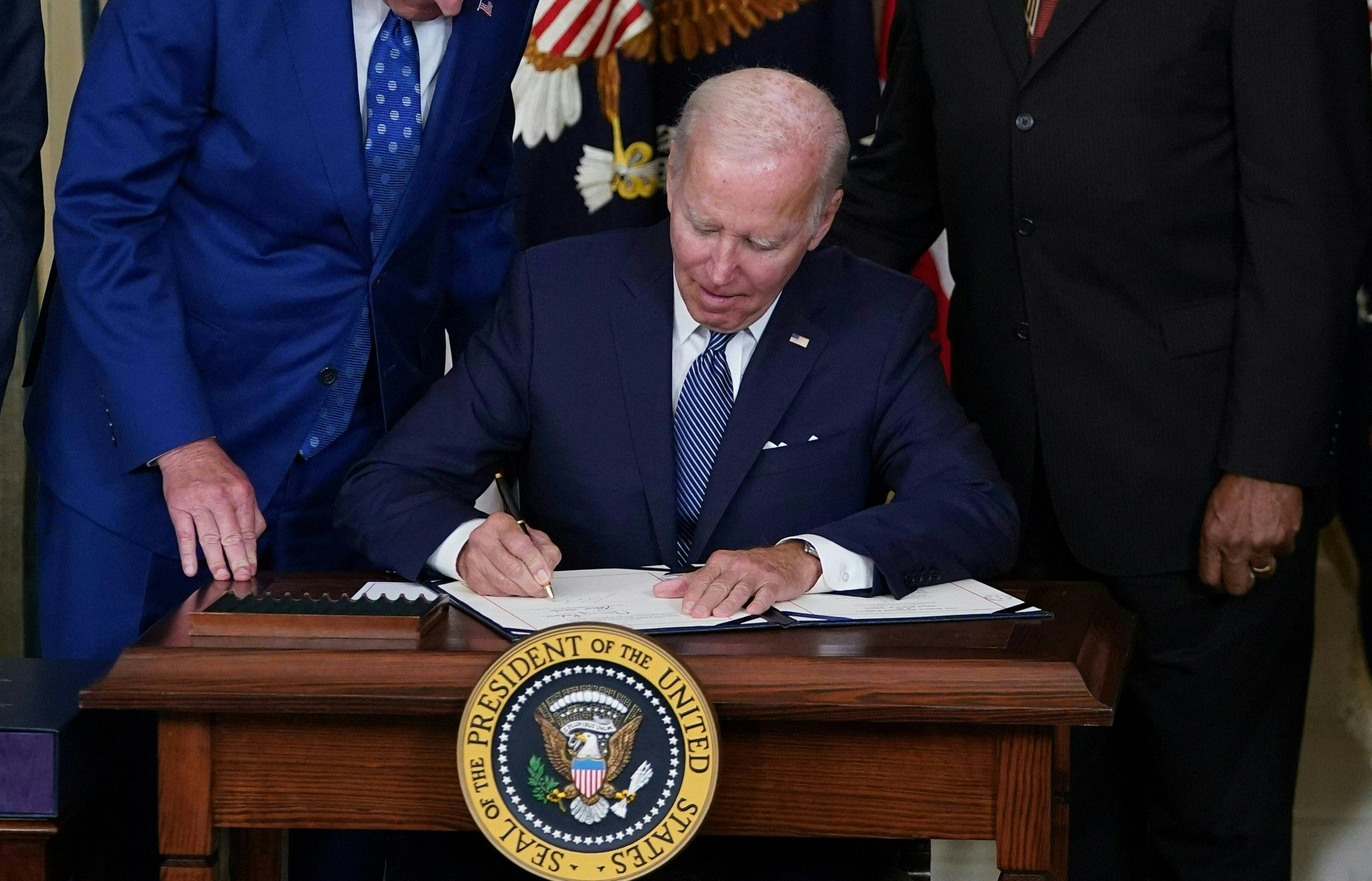 Biden sits at a desk with the presidential seal and signs a bill.