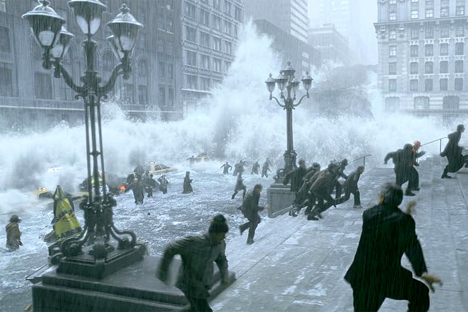 A wave hurtles down a street with tall buildings as people run in all directions. 