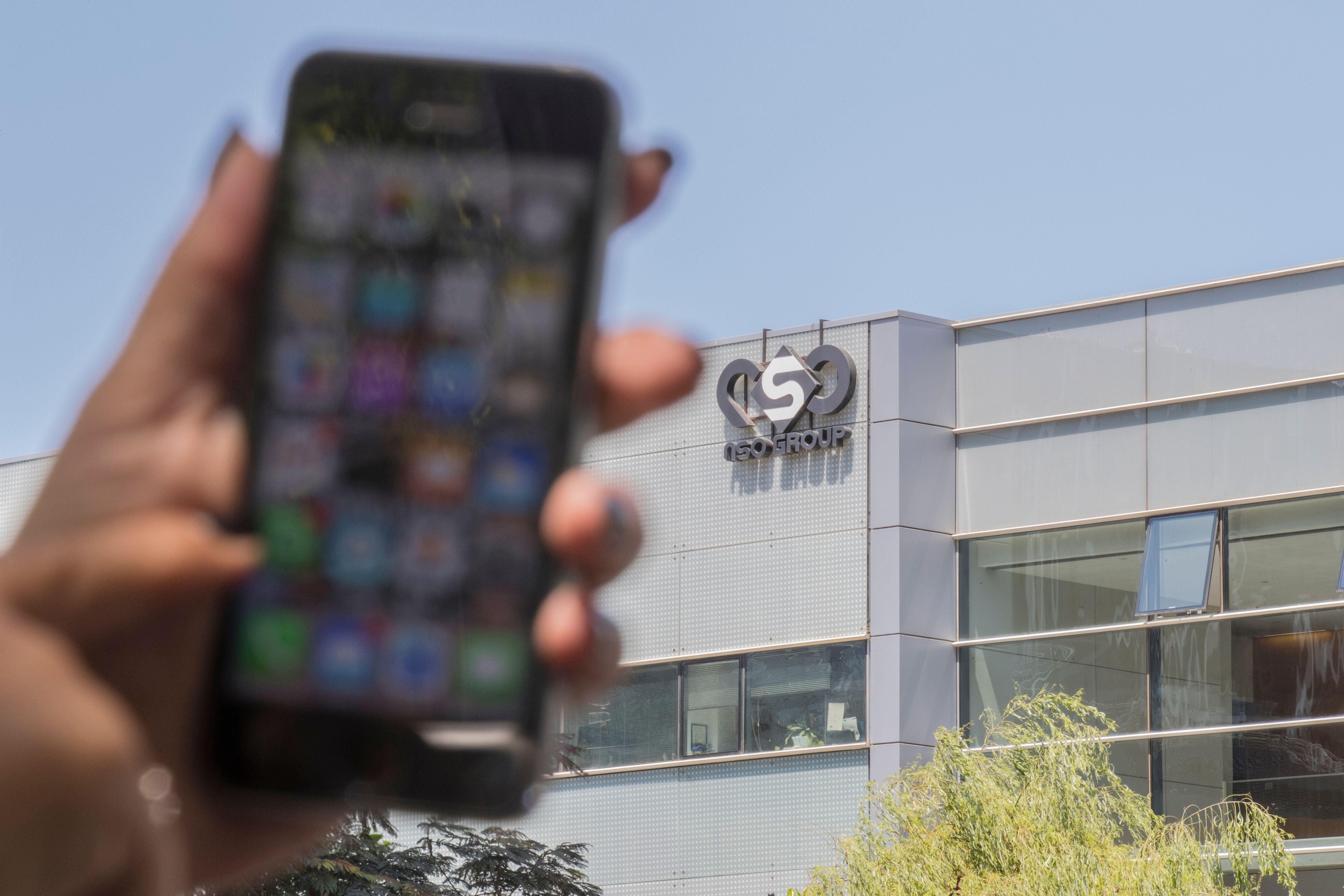 A woman holds a blurry iPhone in front of a large gray office building with the NSO Group logo.