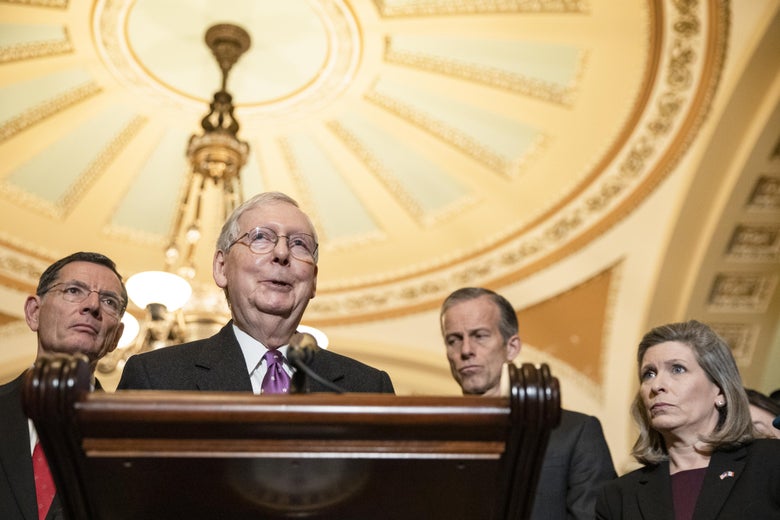 Mitch McConnell speaks at a lectern, surrounded by people standing behind him.