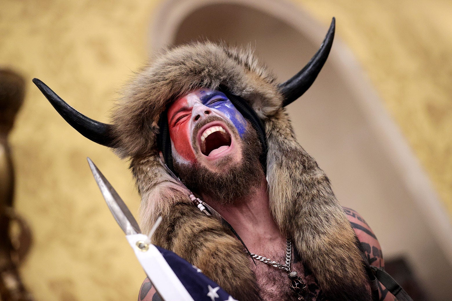 A shirtless, screaming man wearing a raccoon hat with horn screams in the Capitol.
