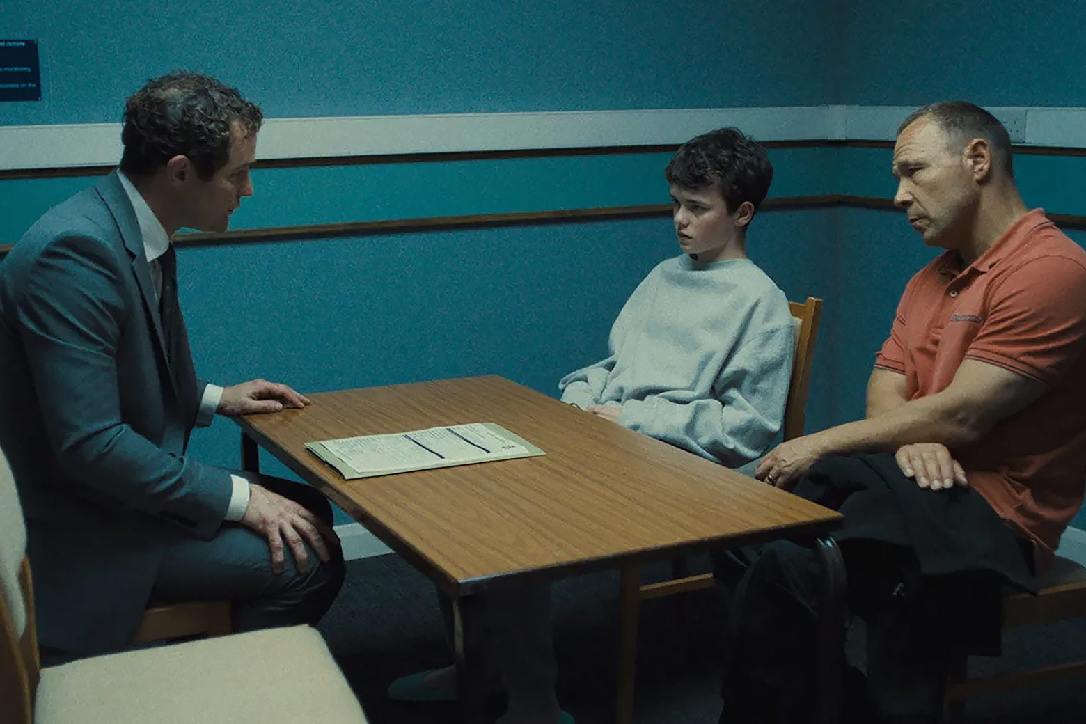 A young boy in a gray sweatshirt sits at a table in an interrogation room with his father as he is questioned by a detective.