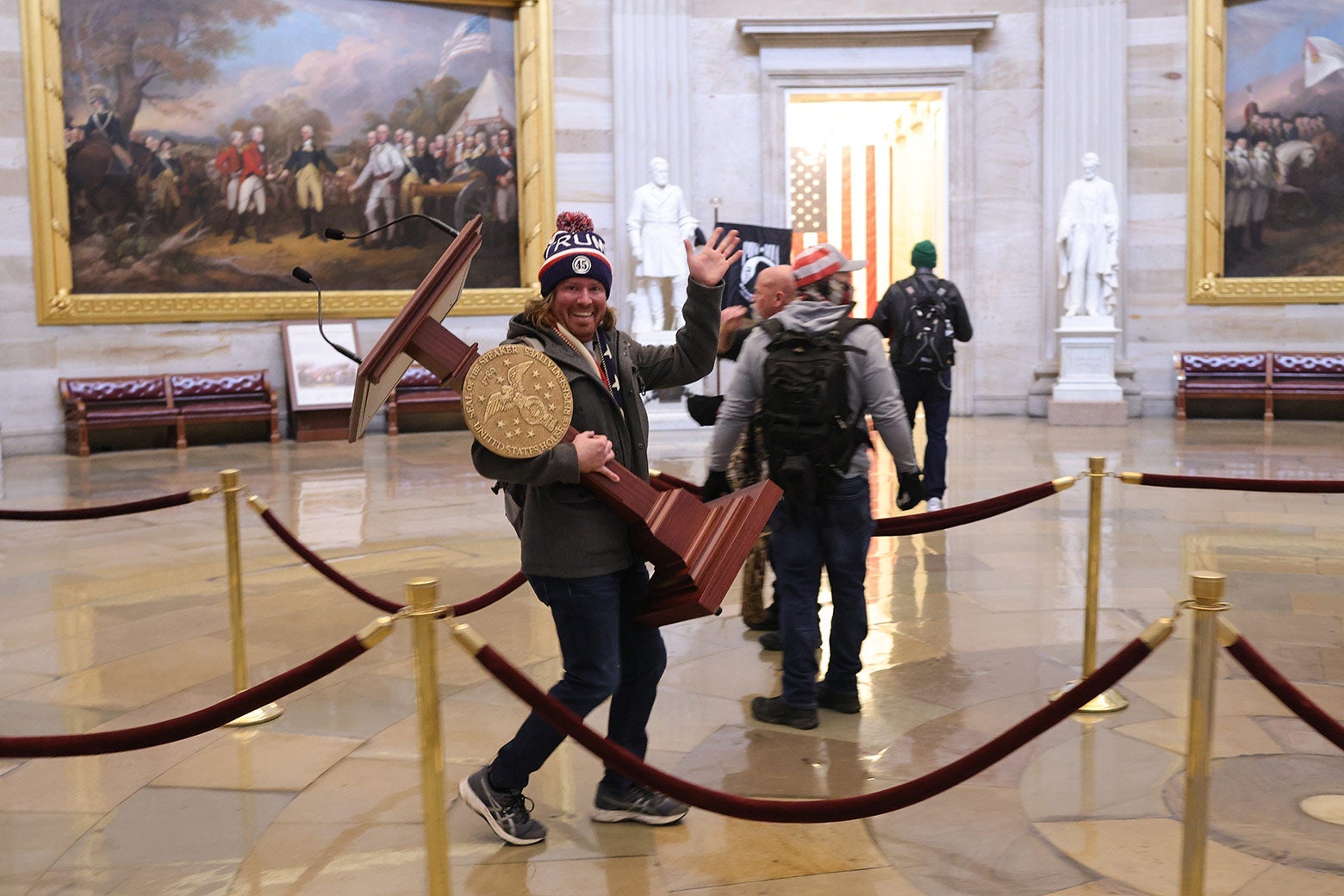 A protester in a Trump beanie smiles and waves at the camera while walking through the Capitol holding a podium for the speaker of the House.