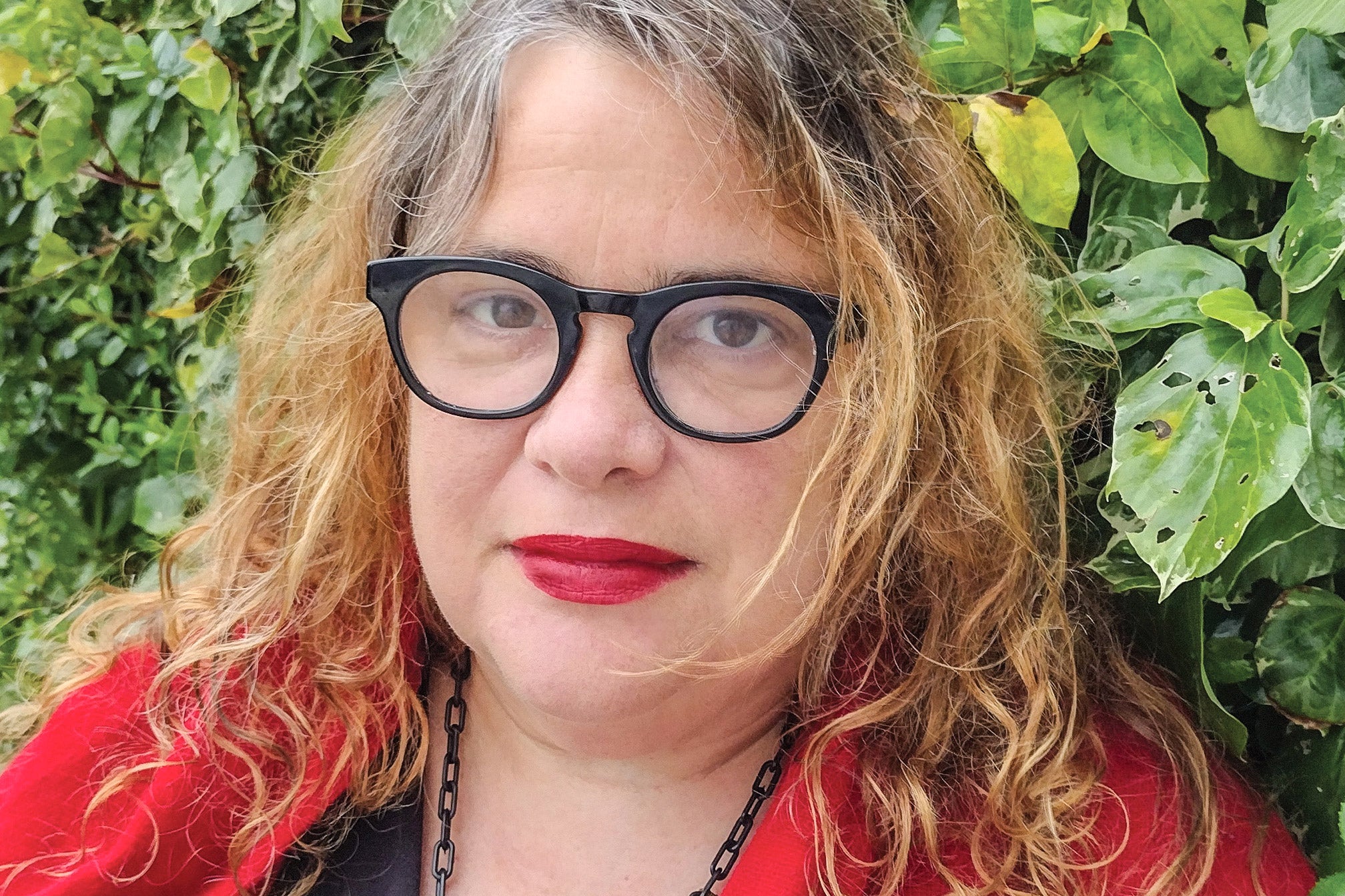 A white woman with wavy brown hair and glasses and red lipstick looks at the camera.