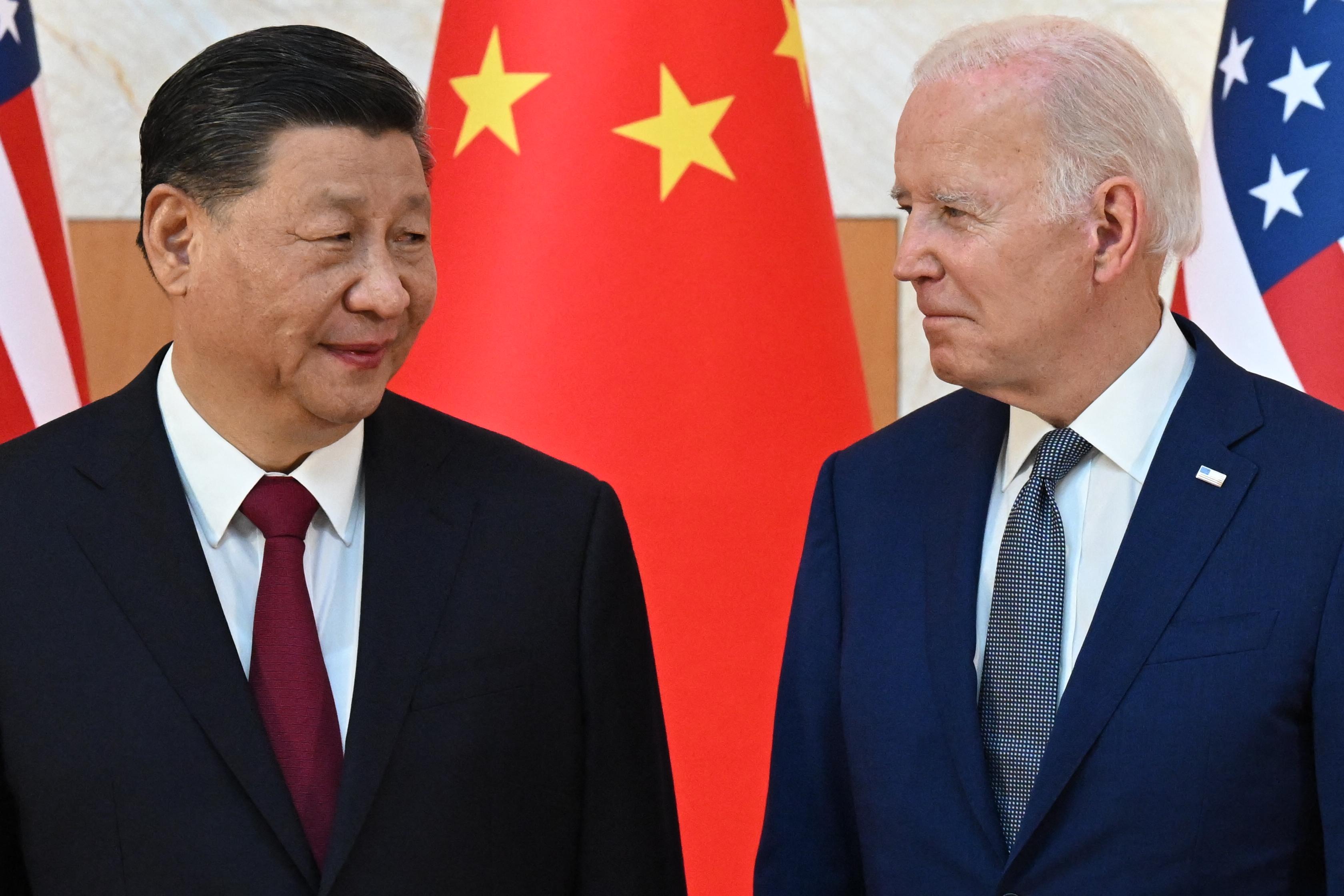 Chinese President Xi Jinping and U.S. President Joe Biden standing side by side and looking at each other, with Chinese and American flags in the background.