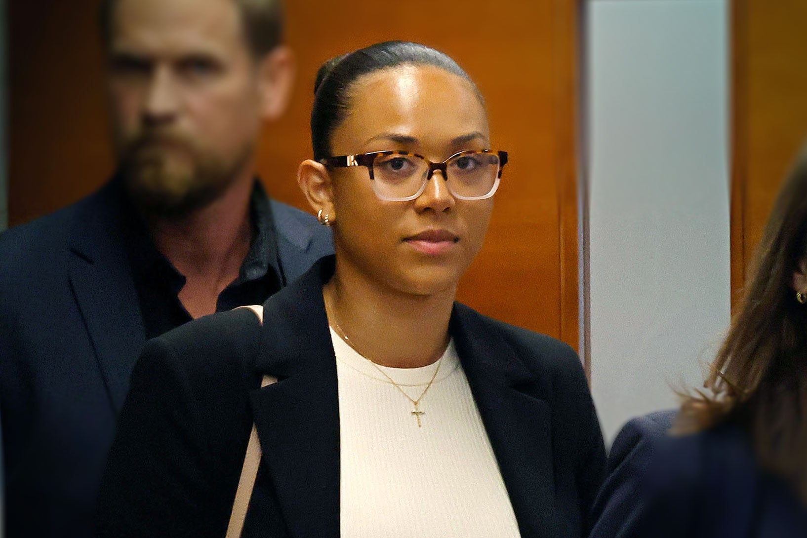 A woman walking, with her hair pulled back in a sleek bun, and wearing tortoiseshell glasses, a white top, a gold cross necklace, and a dark blazer.