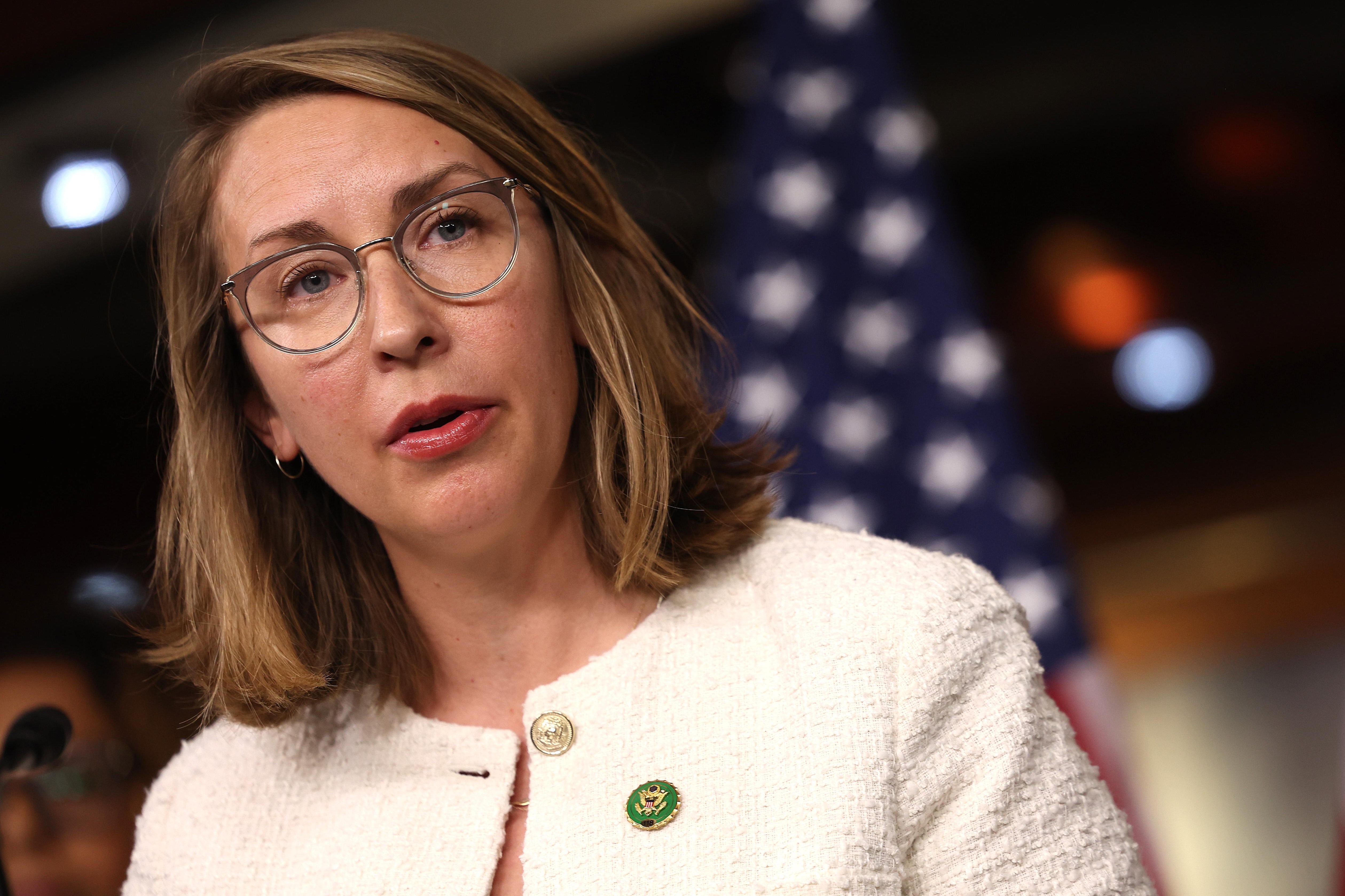 A white woman with round glasses and a white jacket on speaks.