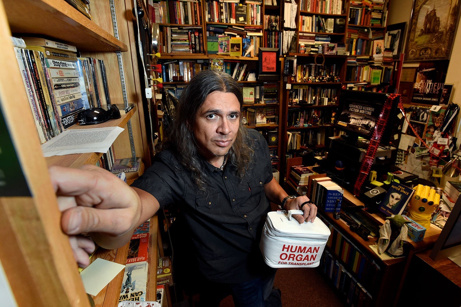 Stephen Graham Jones stands in a room full of books, carrying a box labeled "Human Organ."