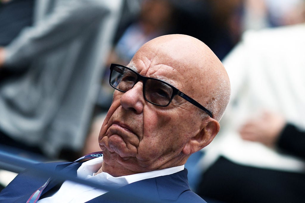 Murdoch, seen at a tilted angle in the stands at a tennis match, stares forward with a grumpy expression.