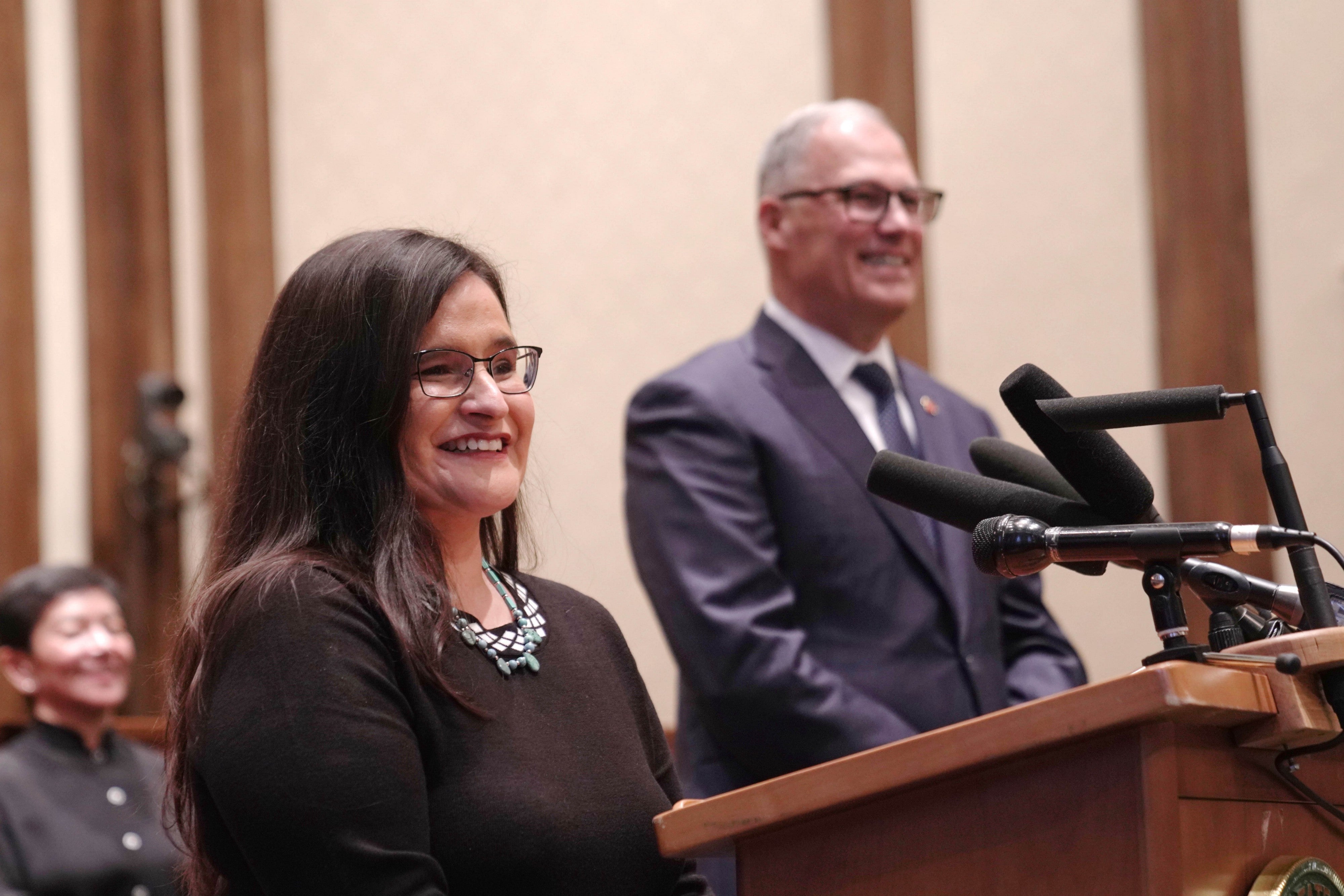 Raquel Montoya-Lewis smiles, standing at a podium next to Jay Inslee as he announces her appointment at a formal ceremony.