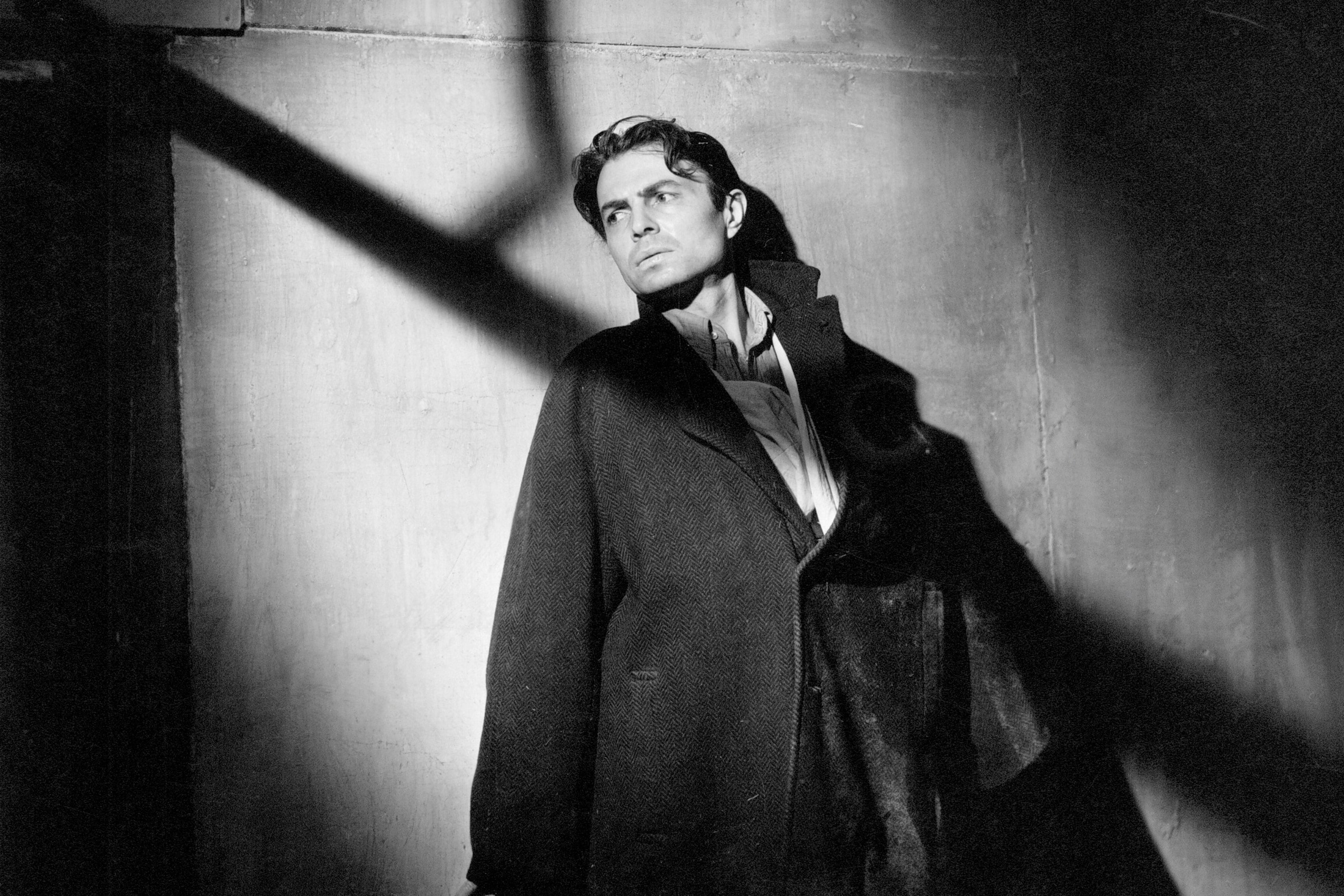 In a black and white image, a man leans against a wall facing a light source from off screen which leave shadows splayed across his face.