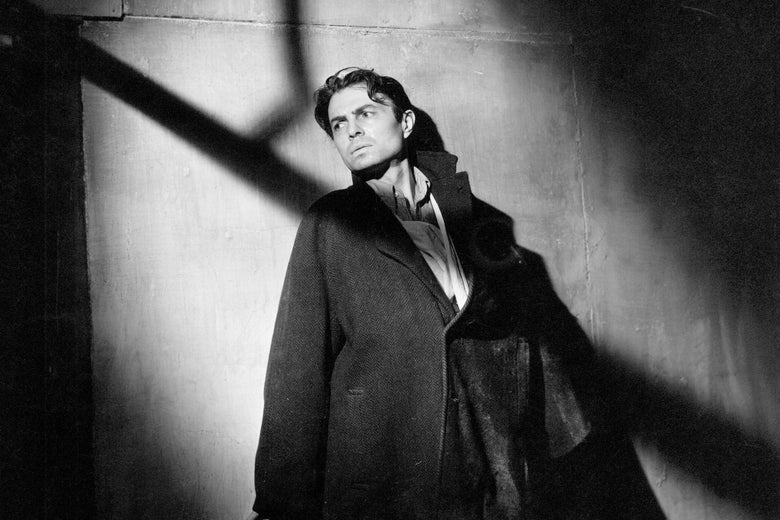 In a black and white image, a man leans against a wall facing a light source from off screen which leave shadows splayed across his face. 