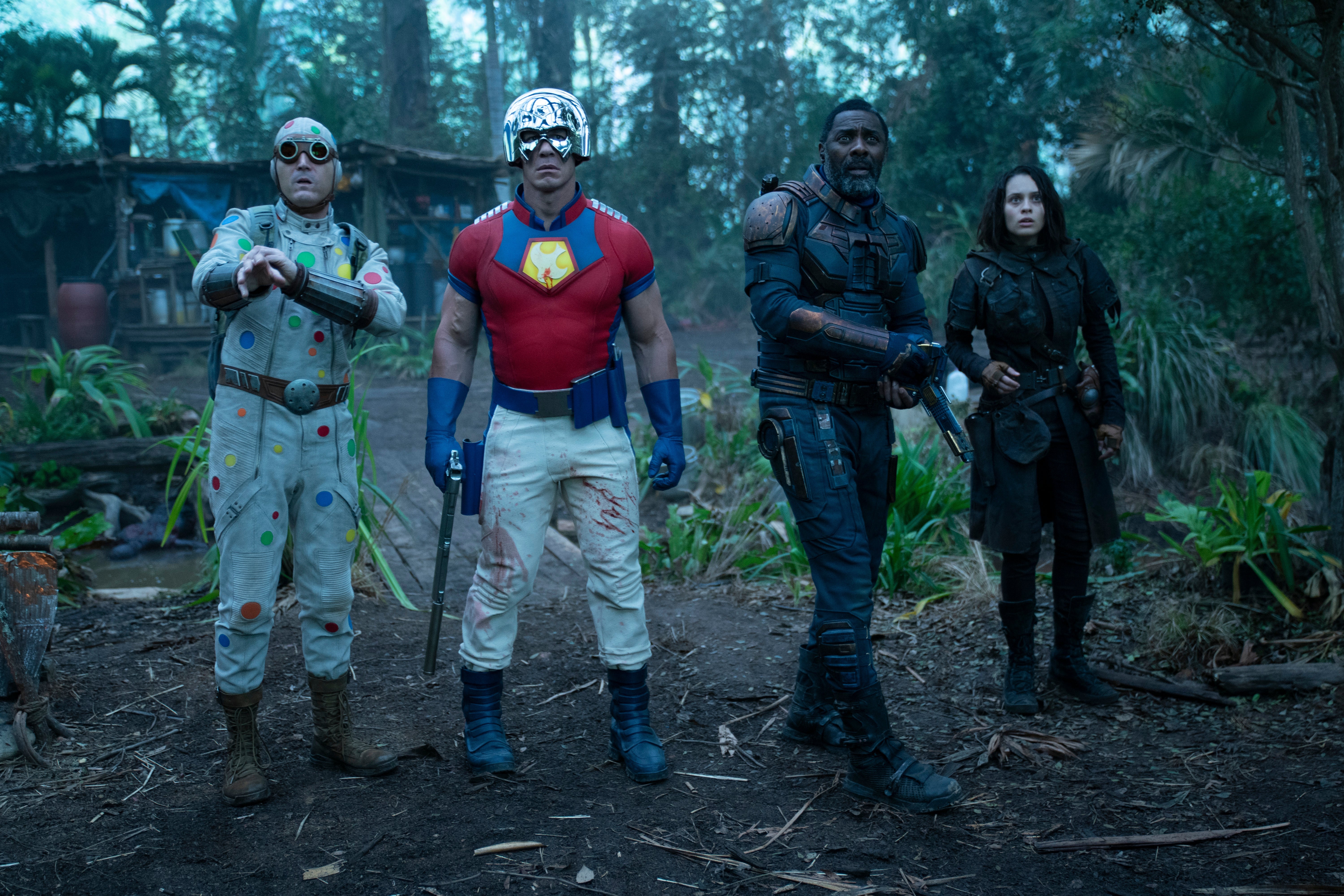 In a forest, a man wearing a crudely made polka dot-covered jumpsuit, a man in a bright red shirt and a metal helmet, and a man and woman both wearing all black. 
