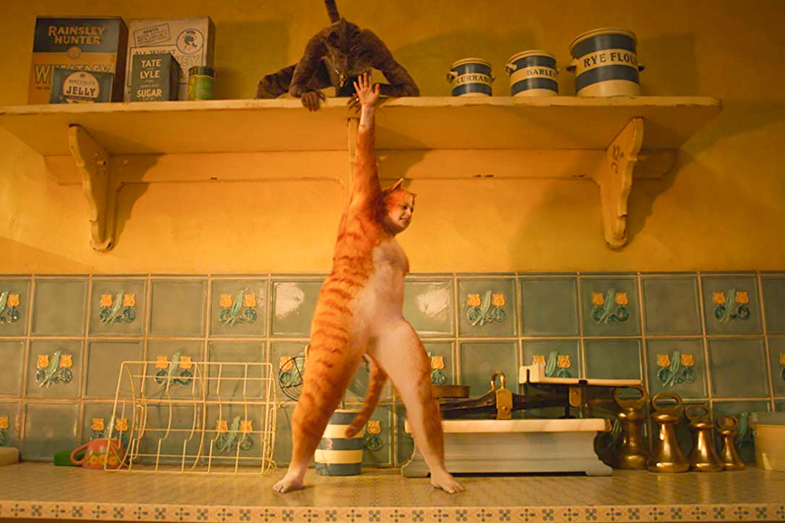 Rebel Wilson, wearing a yellow cat suit with a white belly, stands on a kitchen counter with a floral tile backsplash, one arm reaching up toward a shelf with boxes and jars.