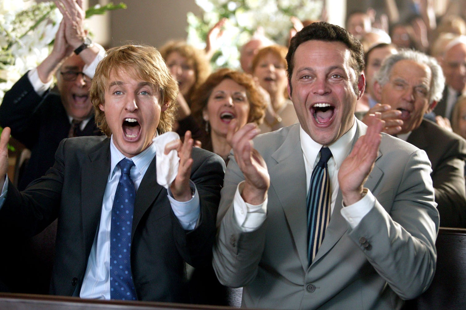 Owen Wilson and Vince Vaughn, in suit jackets and ties in a wedding pew, cheer excitedly.