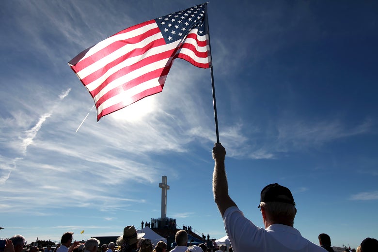 A man, seen from behind, holds up an American flag. He is standing in a crowd of people all facing a large cross.