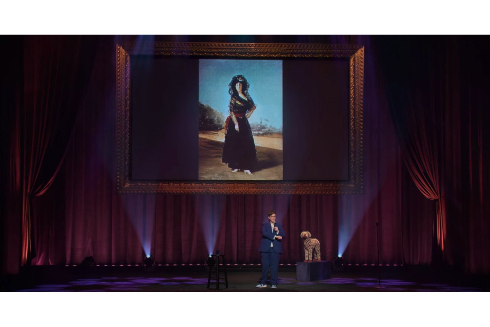 Hannah Gadsby onstage in front of a projection of a Goya painting of a woman in a black dress and white shoes.
