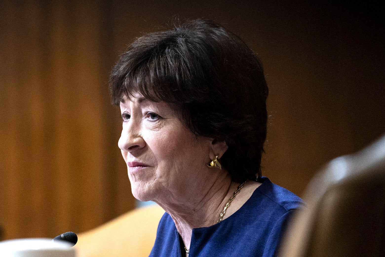 Susan Collins sits in a blue top, behind a set of microphones, gazing to the left.