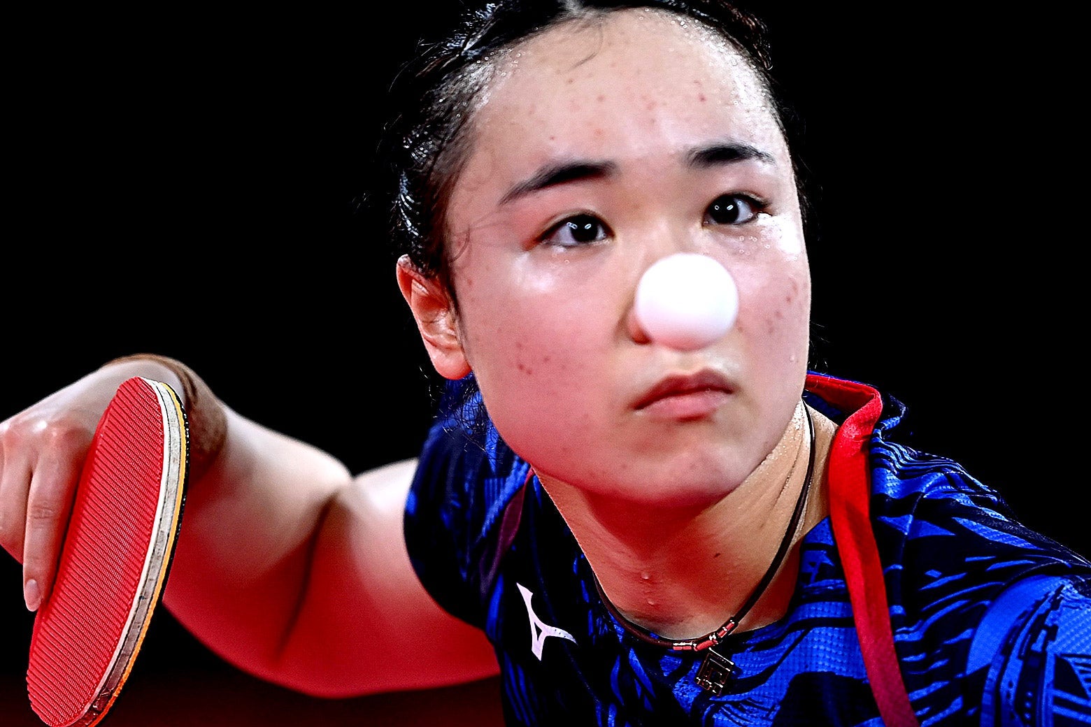 A table tennis player stares intently as she holds her paddle. The white ping pong ball is centered on her face like a clown nose.