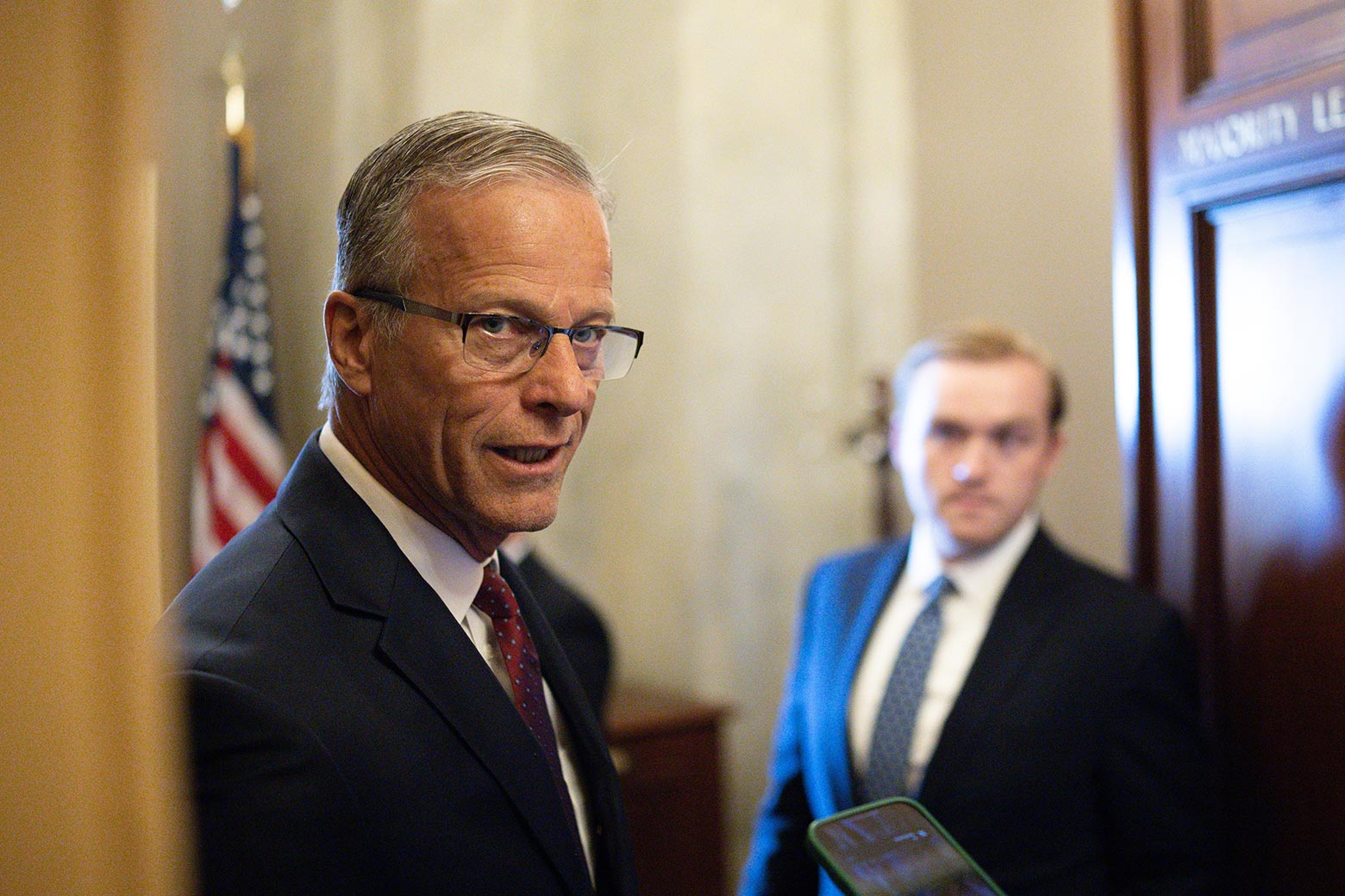An older white man wears glasses and speaks through a door to a reporter as an aide looks on in the background.