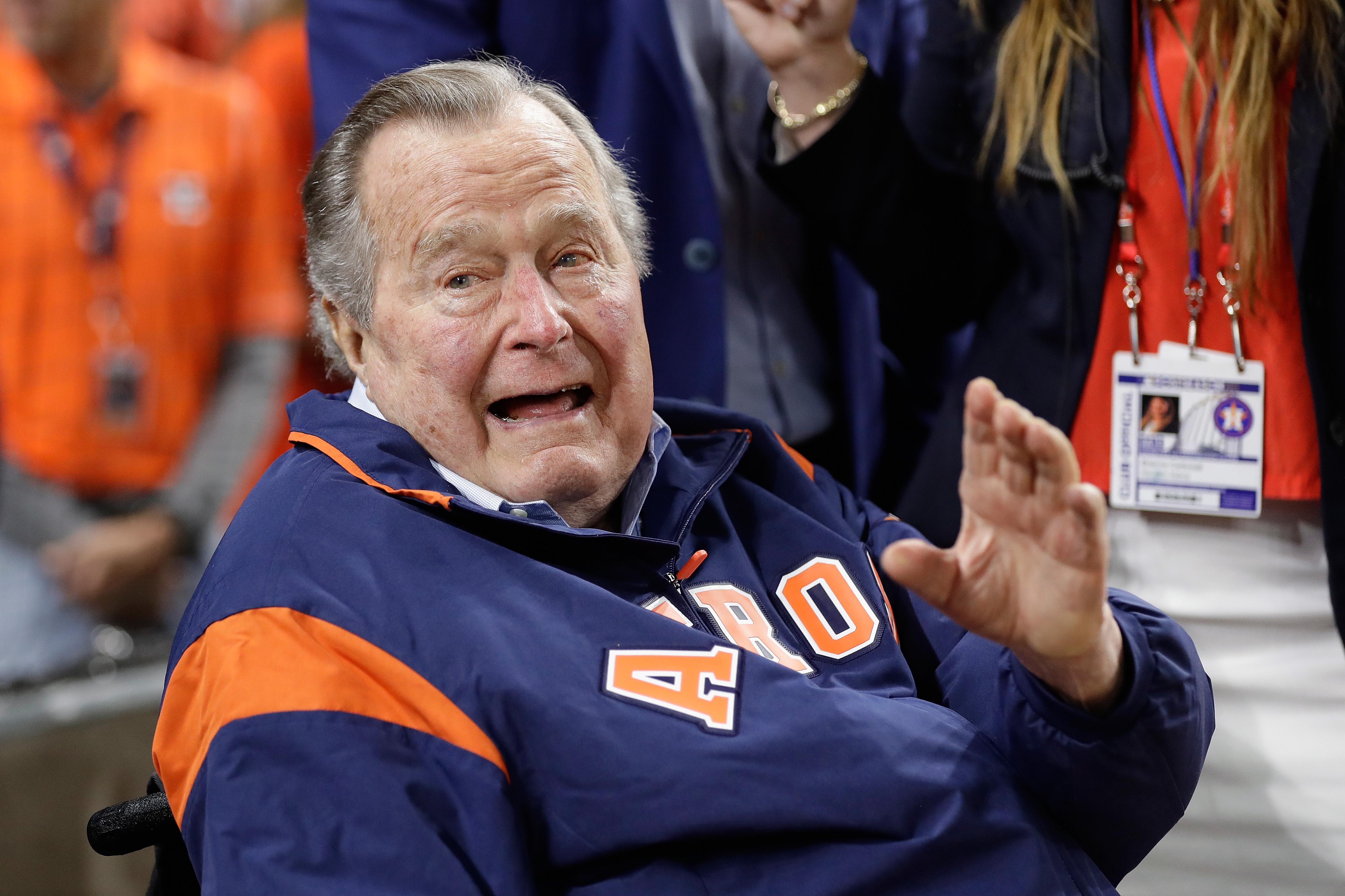 George H. W. Bush waves to the camera. He is wearing an Astros jacket, and figures in sports-related attire can be seen behind him.