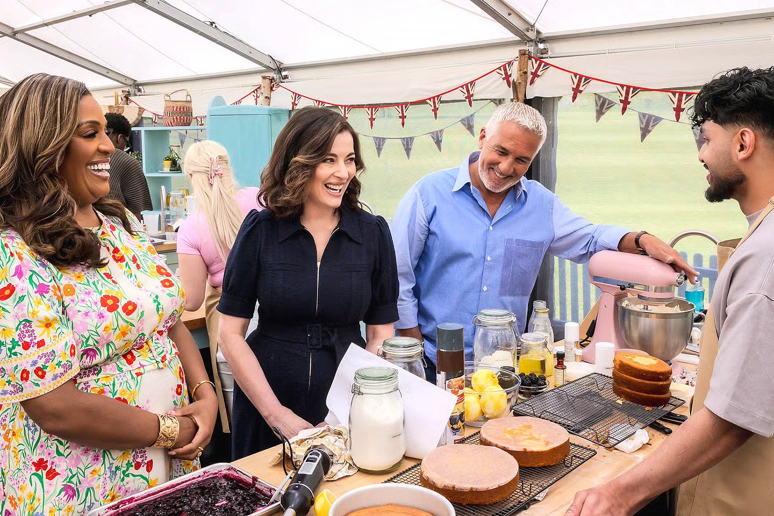 Three judges stand at a counter ready to judge a cake on the Great British Bake-Off.