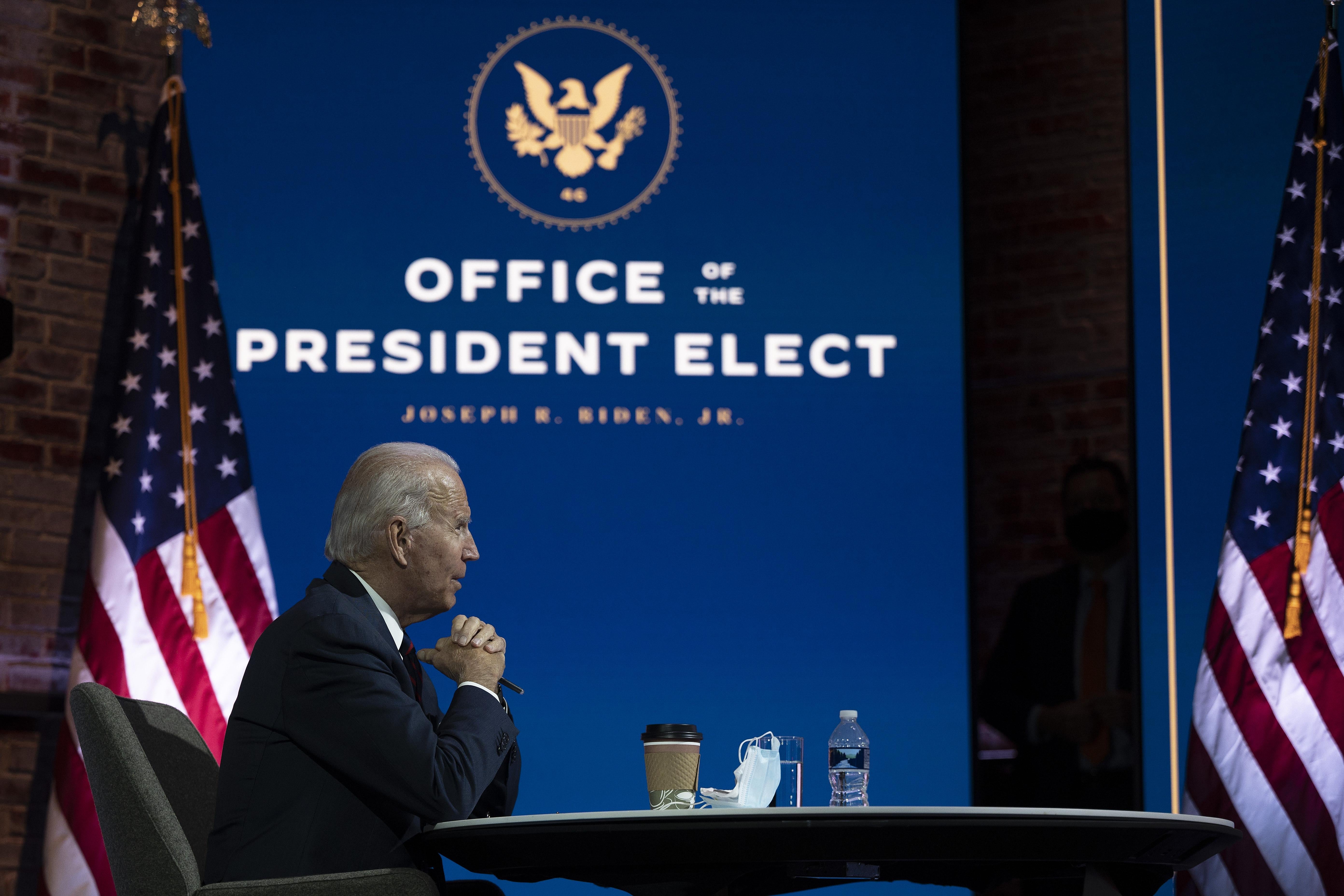 Joe Biden rests his elbows on a table with text reading "Office of the President Elect" behind him.