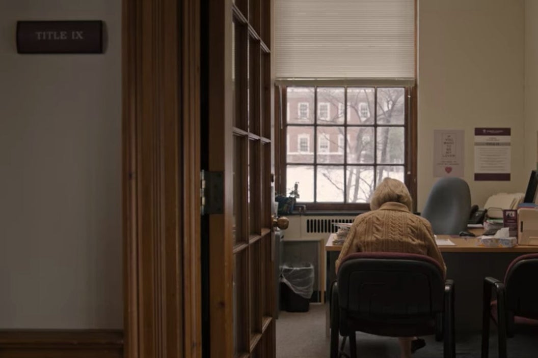 A door opens into an office in a college building, where Joan sits in a chair in front of a desk.