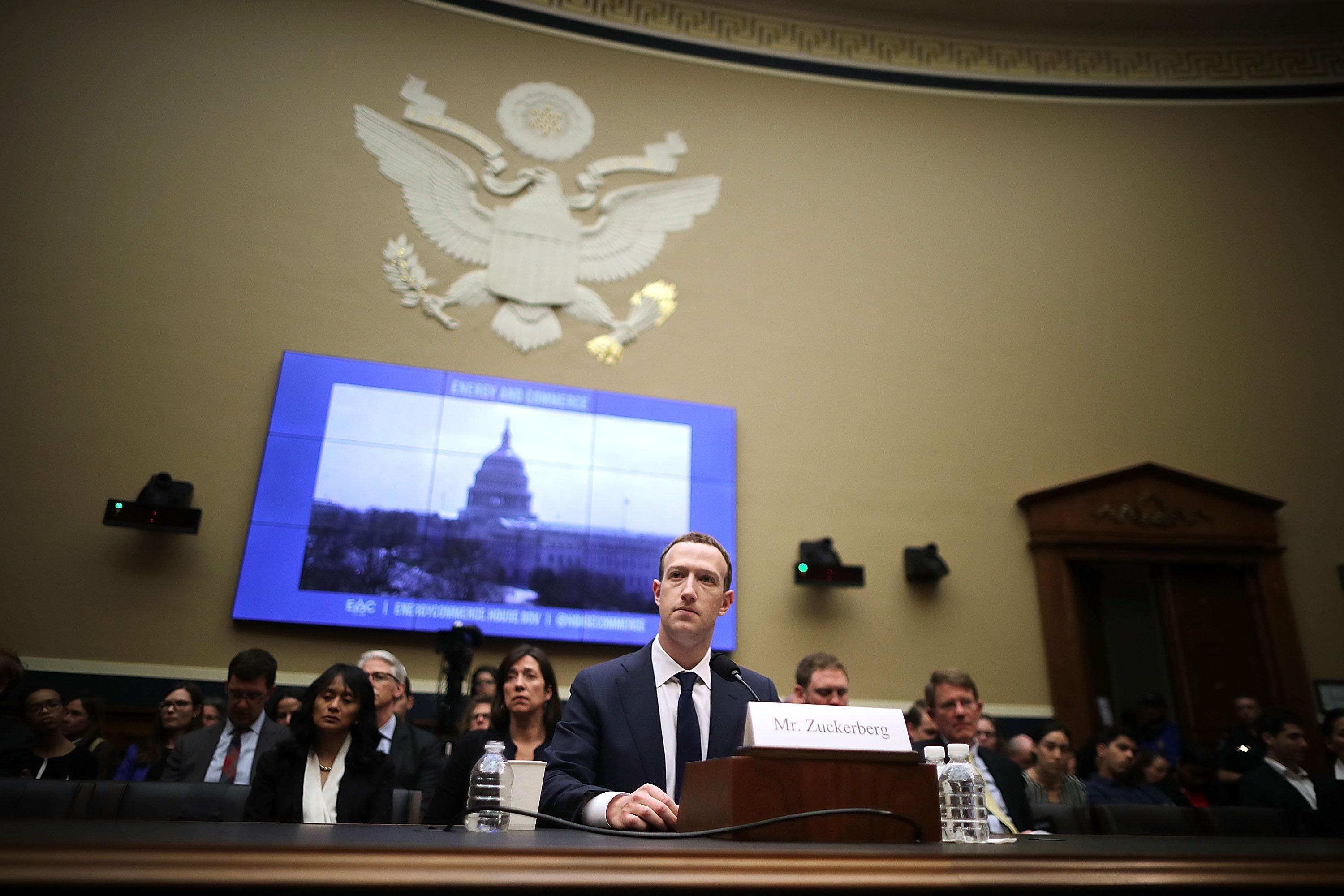 Zuckerberg sits at a table in a large room. Behind him are an audience of watchers, a screen showing an image of the Capital Building, and the Great Seal of the U.S.