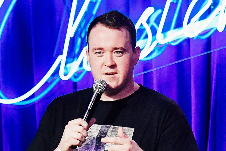 Shane Gillis holds a microphone in front of a neon sign.