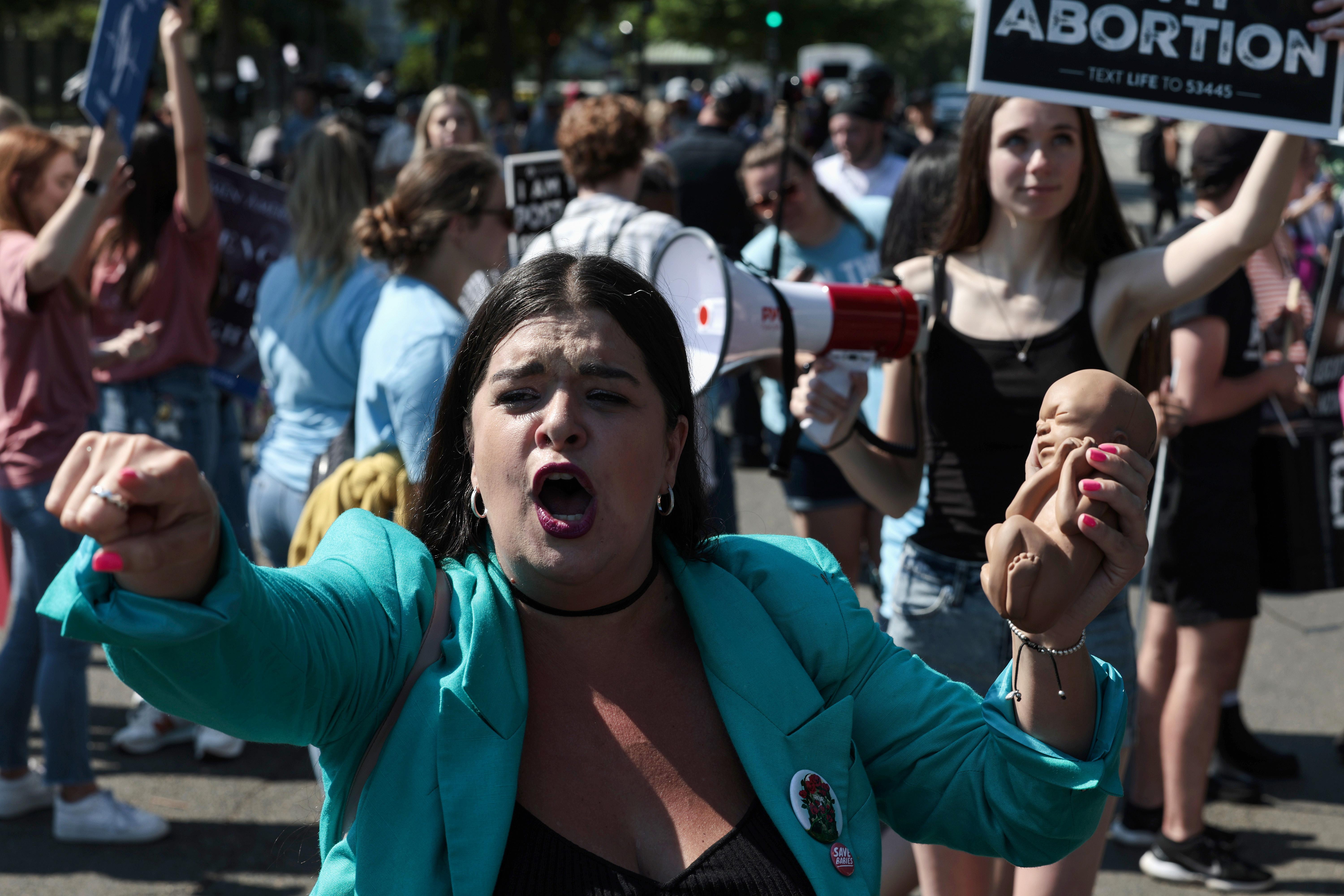 Woman pointing and shouting and holding a plastic doll in front of a crowd.
