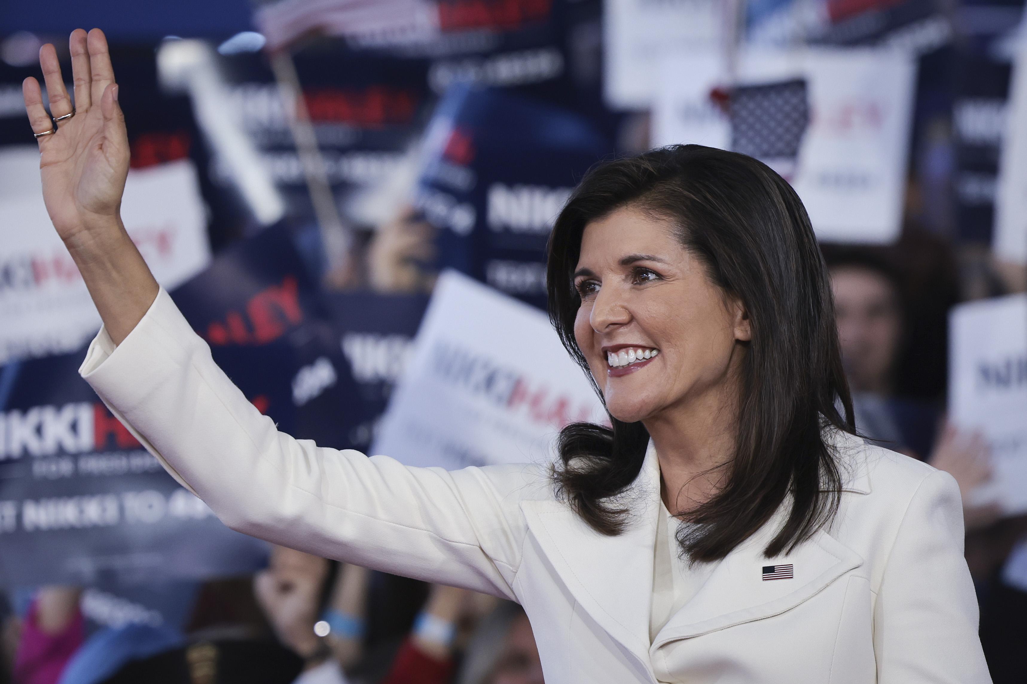 Nikki Haley wavs at supporters while wearing a white jacket with a U.S. flag pin.