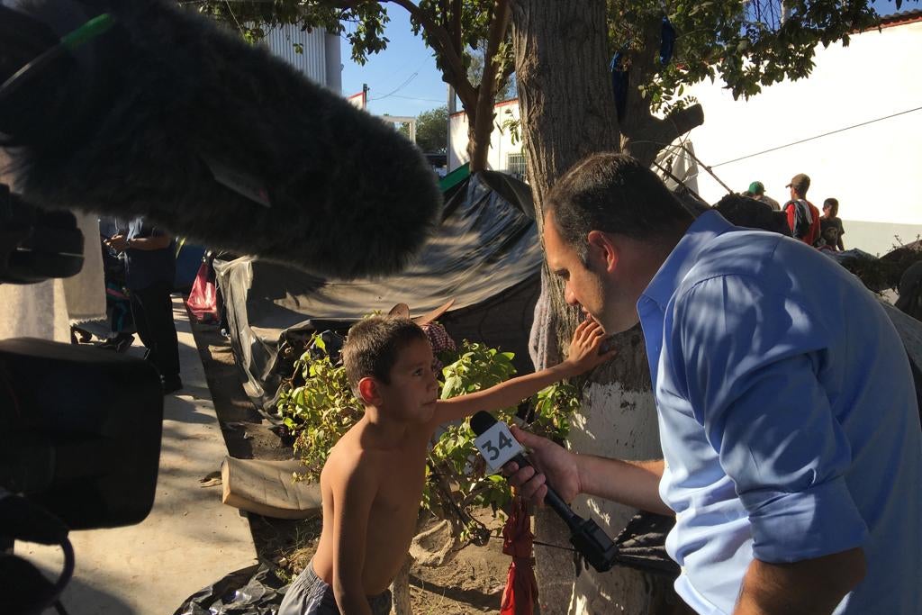 The reporter holds a microphone up to a child leaning against a tree. In the background, a row of makeshift tents in the Tijuana migrant camp.