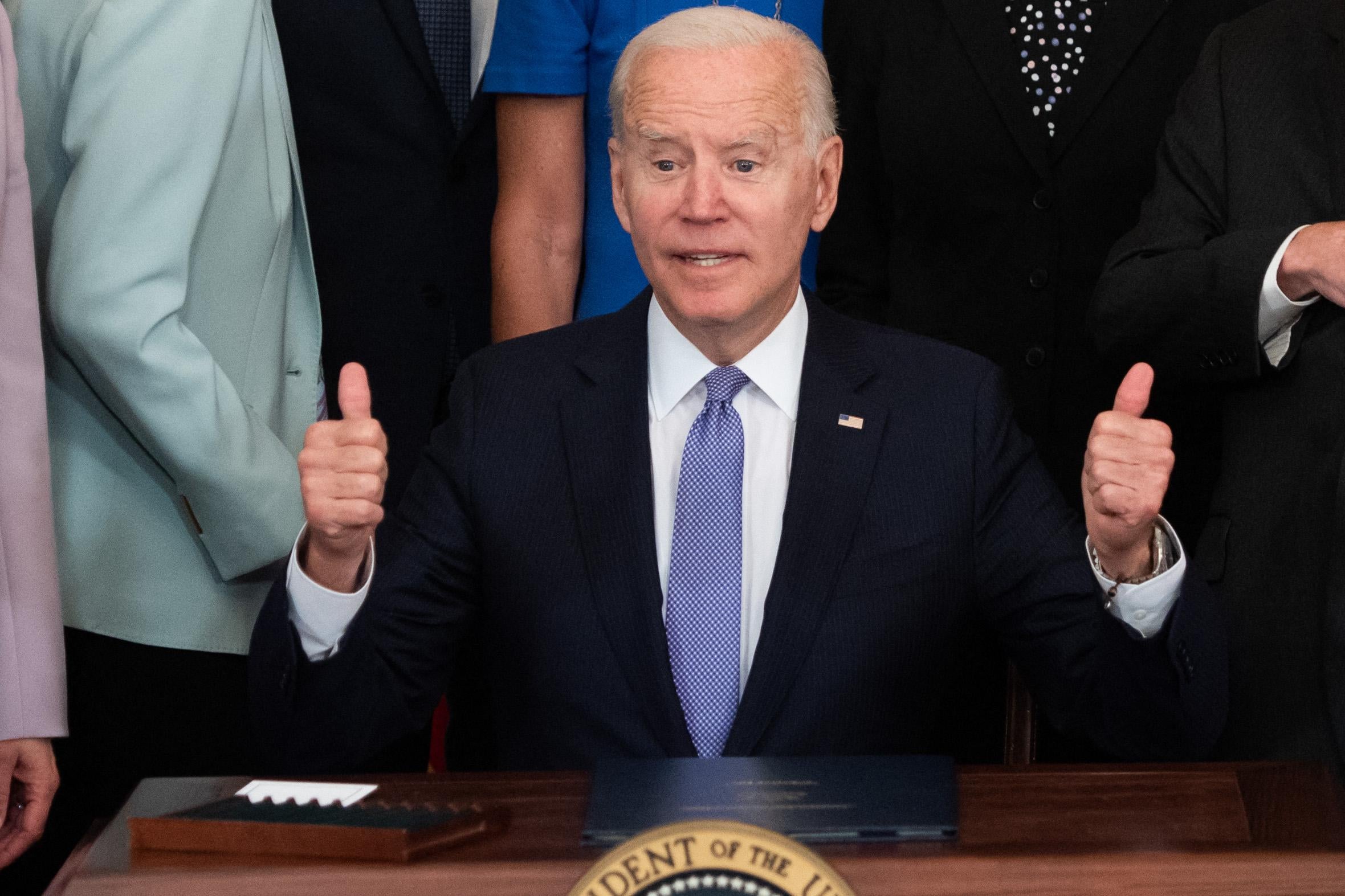 Joe Biden sits at a desk and flashes two thumbs up as people stand behind him.