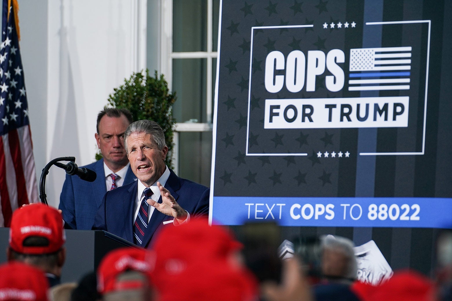 Lynch speaks at a lectern in front of a crowd of people wearing red MAGA hats. A sign to his left says COPS FOR TRUMP.