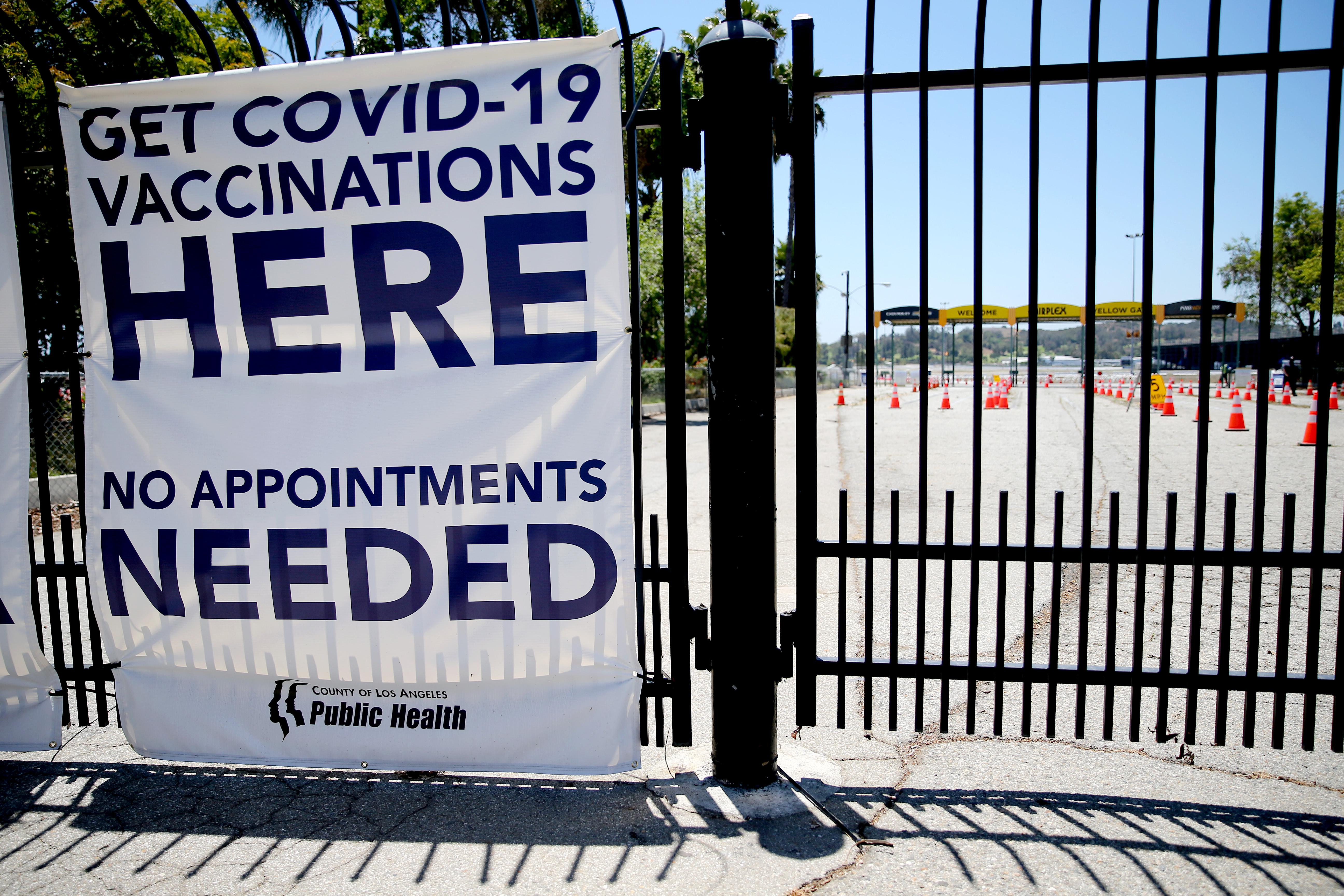 A sign on a black metal gate says "Get COVID-19 Vaccinations Here No Appointments Needed." Behind the gate are rows and rows of empty drive-thru lanes marked by traffic cones.