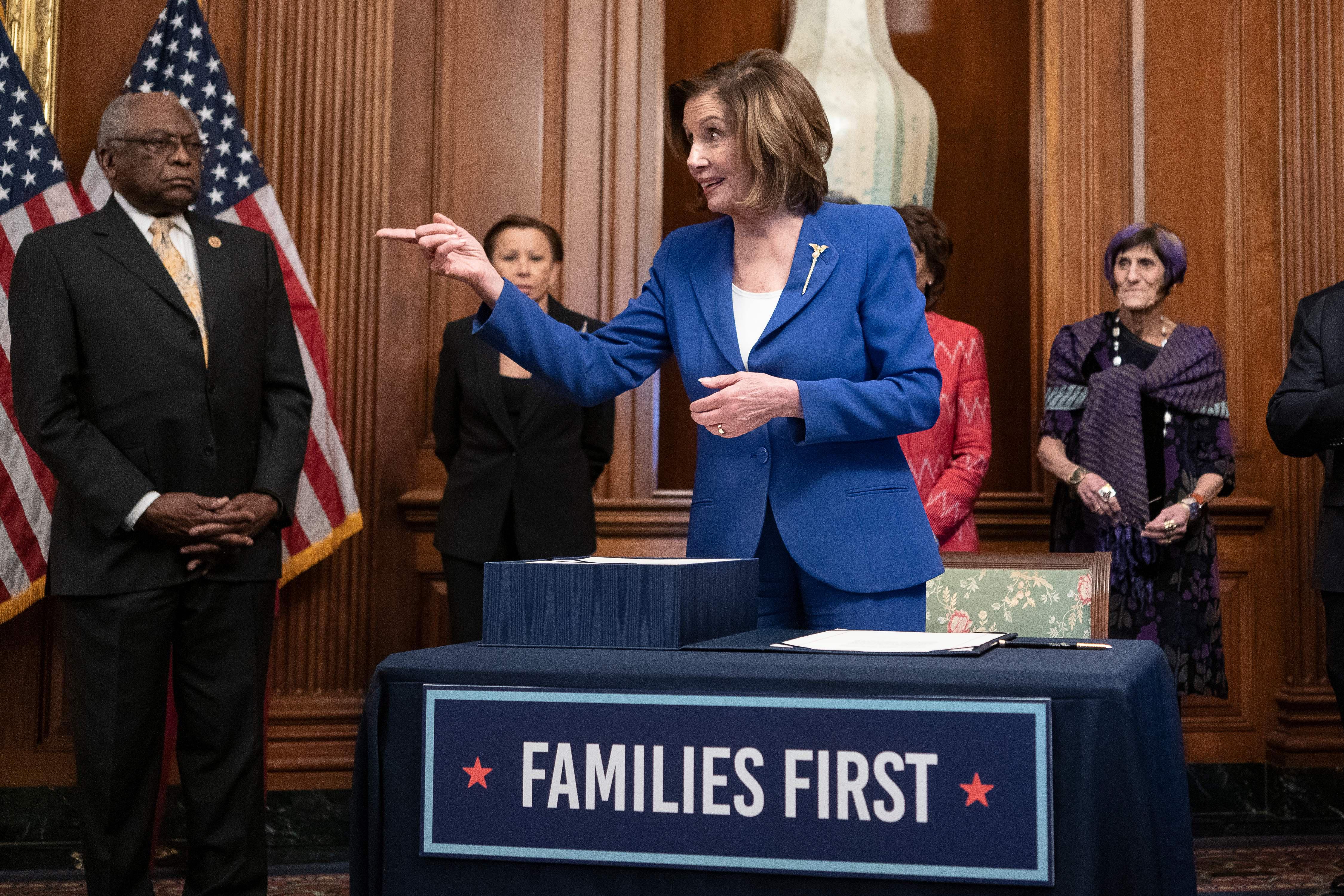 Pelosi points to her right and smiles in front of a table with a banner reading "Families First."