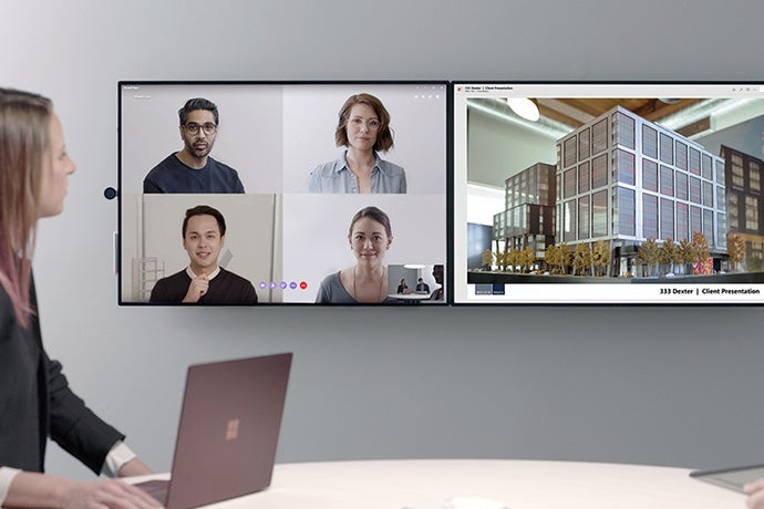 A woman uses the Surface Hub 2 to teleconference with four people as she makes a presentation about a building.